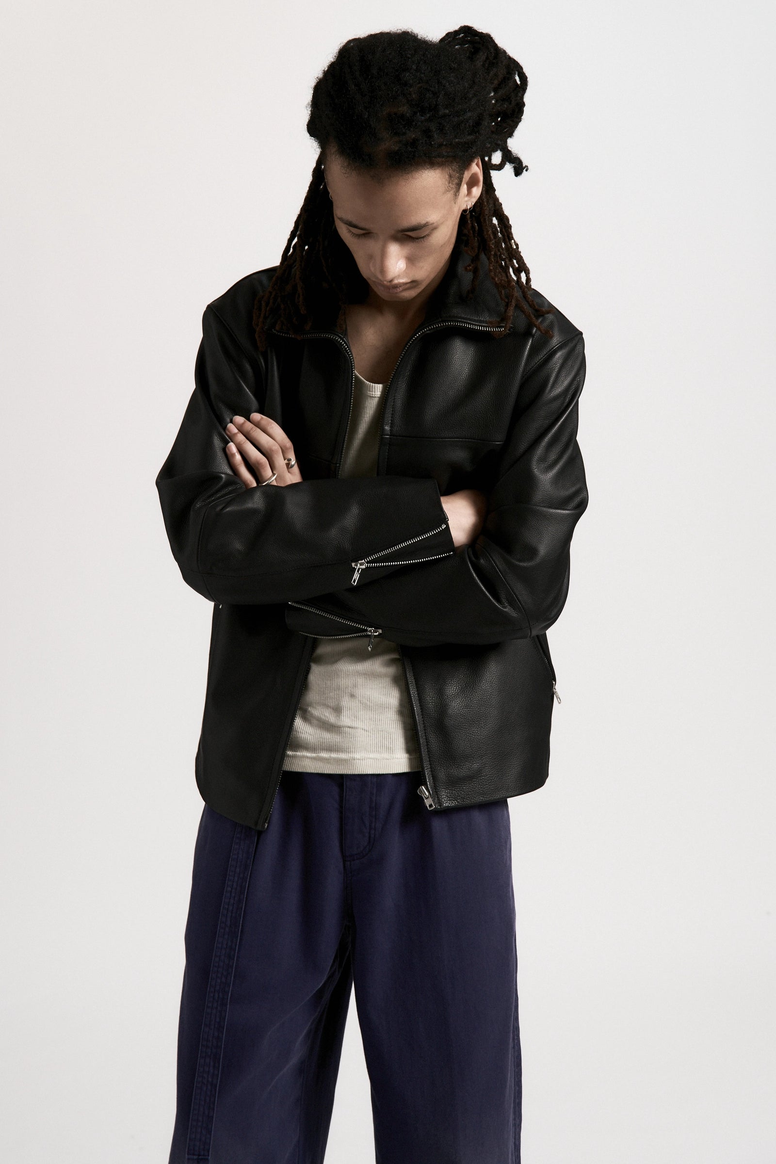 Person looking down with their arms folded wearing a black leather jacket and blue pants on a white background