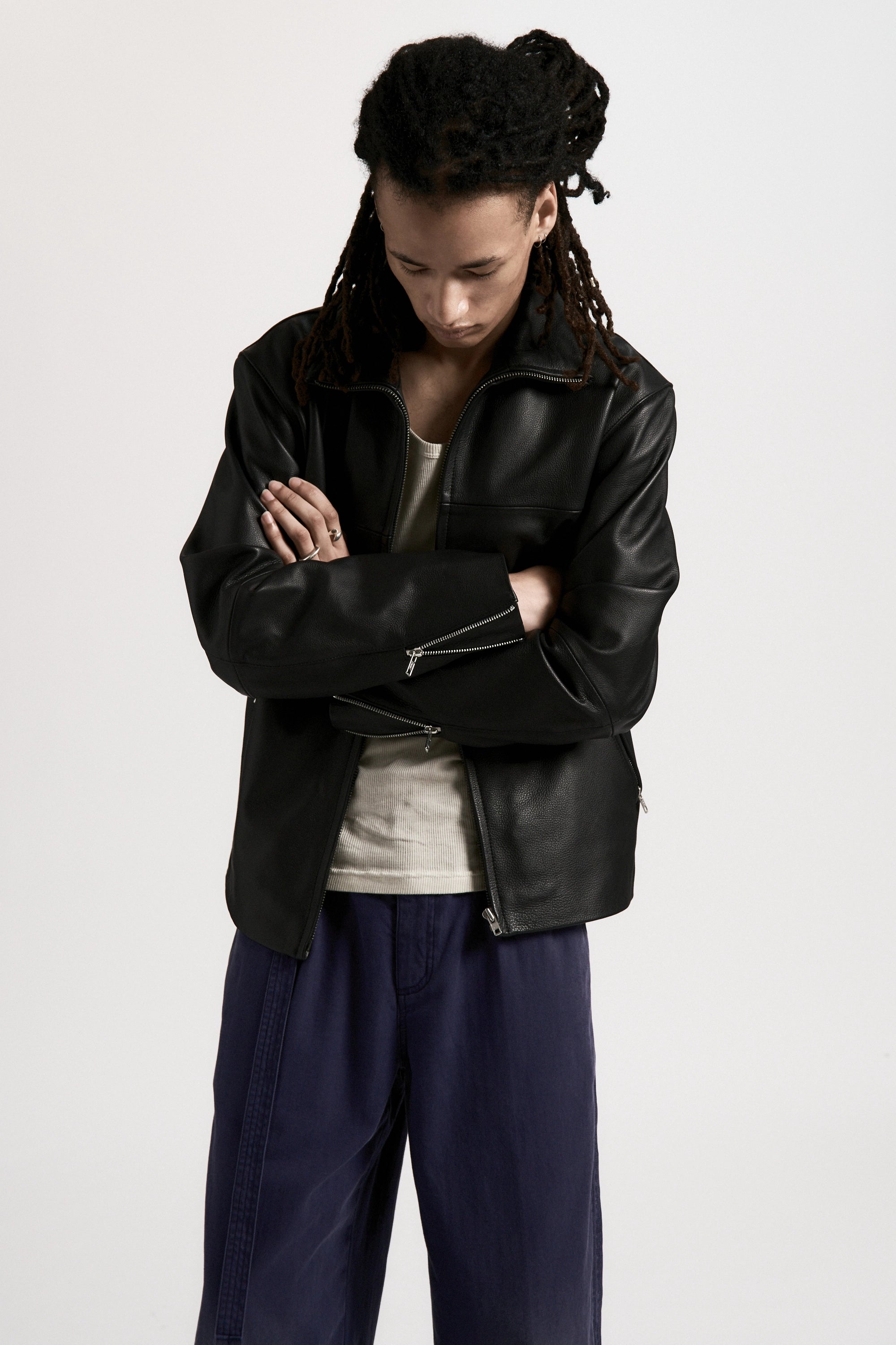 Person looking down with their arms folded wearing a black leather jacket and blue pants on a white background