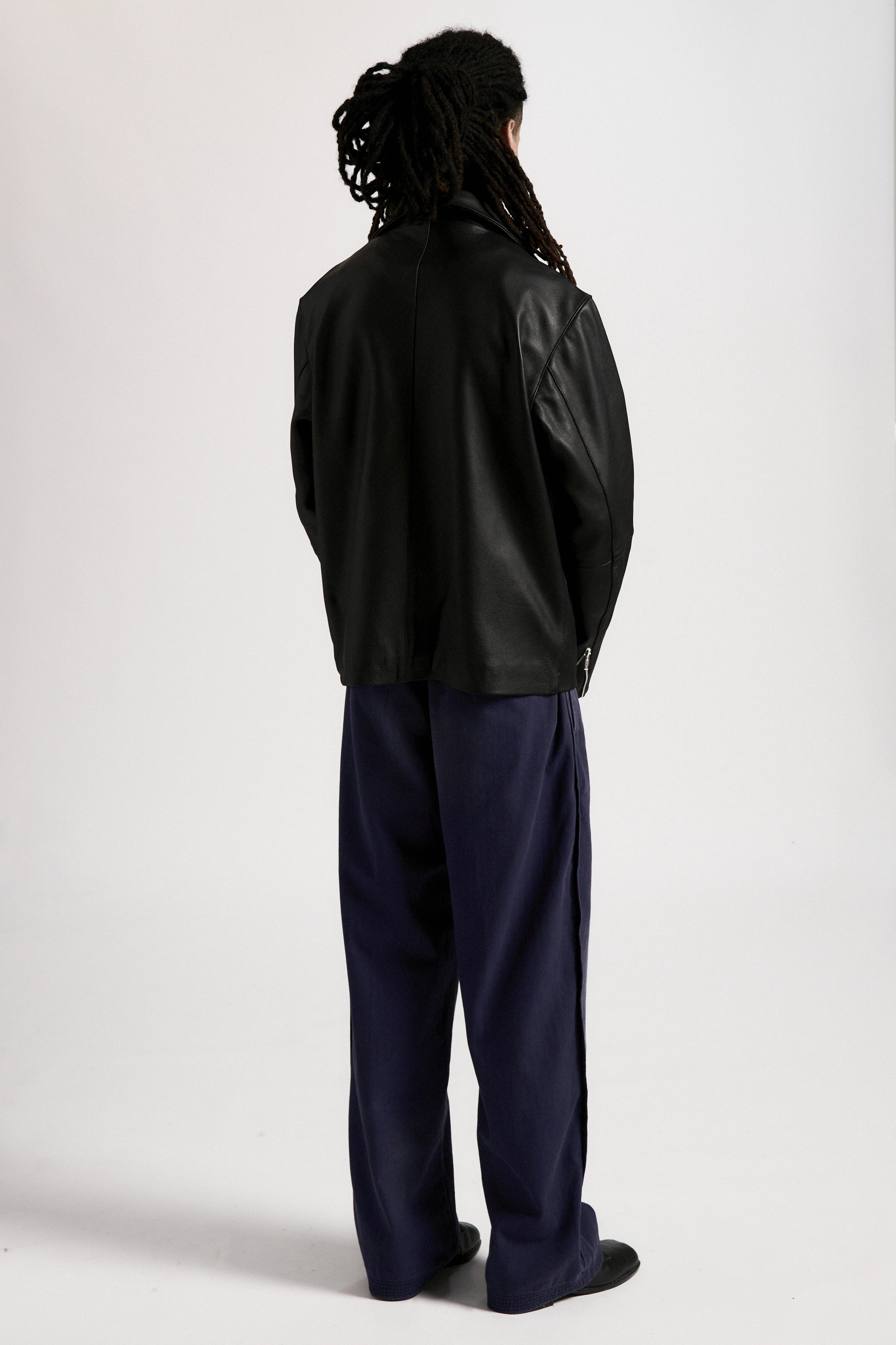 Rear view of a person wearing the men's Bank Jacket made from full grain leather and dark pants on a white background