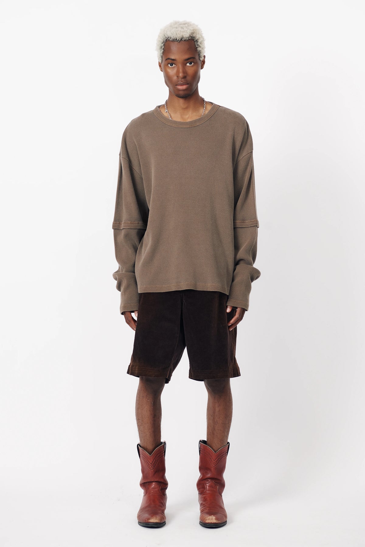 A man standing against a white background wearing washed brown corduroy baggy shorts and a matching long-sleeve sweater, along with brown boots.