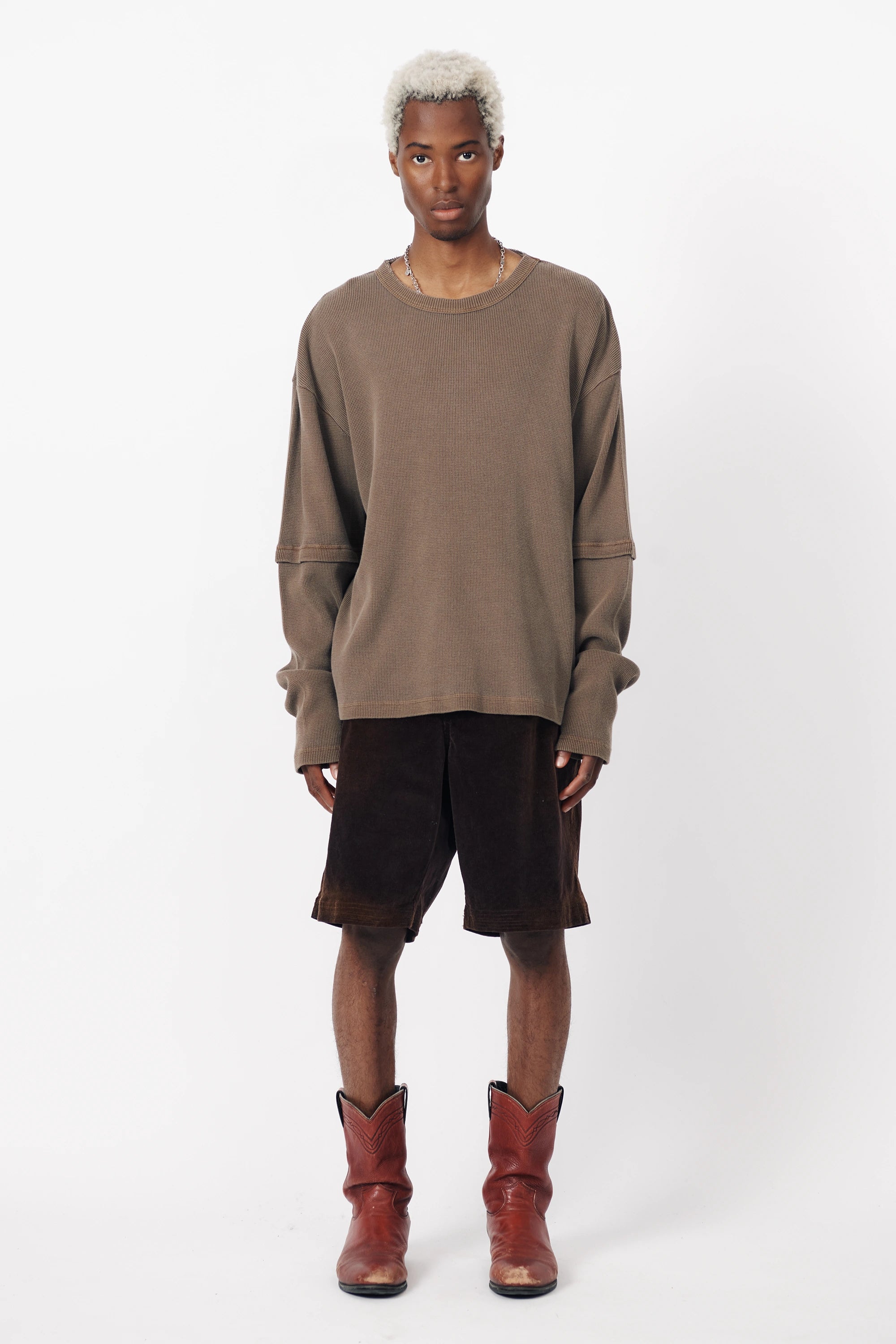 A man standing against a white background wearing washed brown corduroy baggy shorts and a matching long-sleeve sweater, along with brown boots.