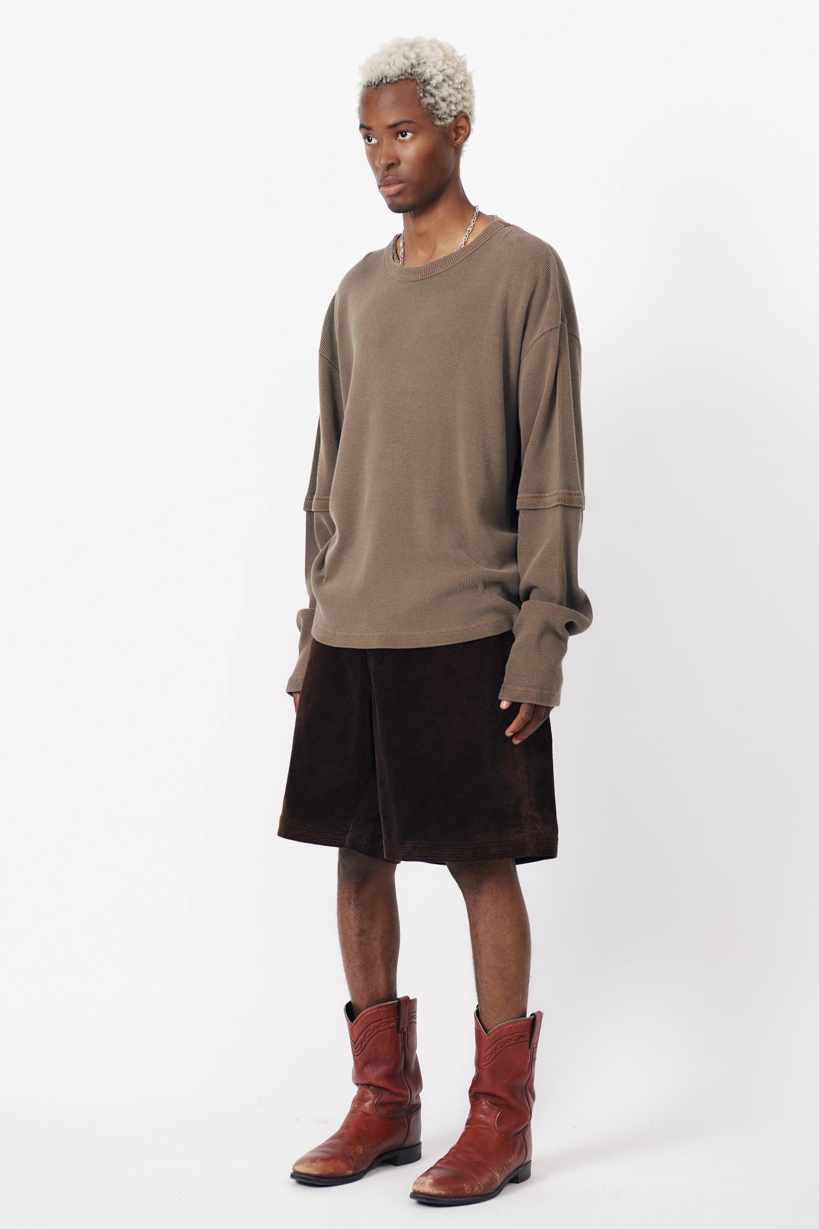 A person standing against a white background wearing a washed brown long-sleeve shirt and dark baggy shorts, with red boots.