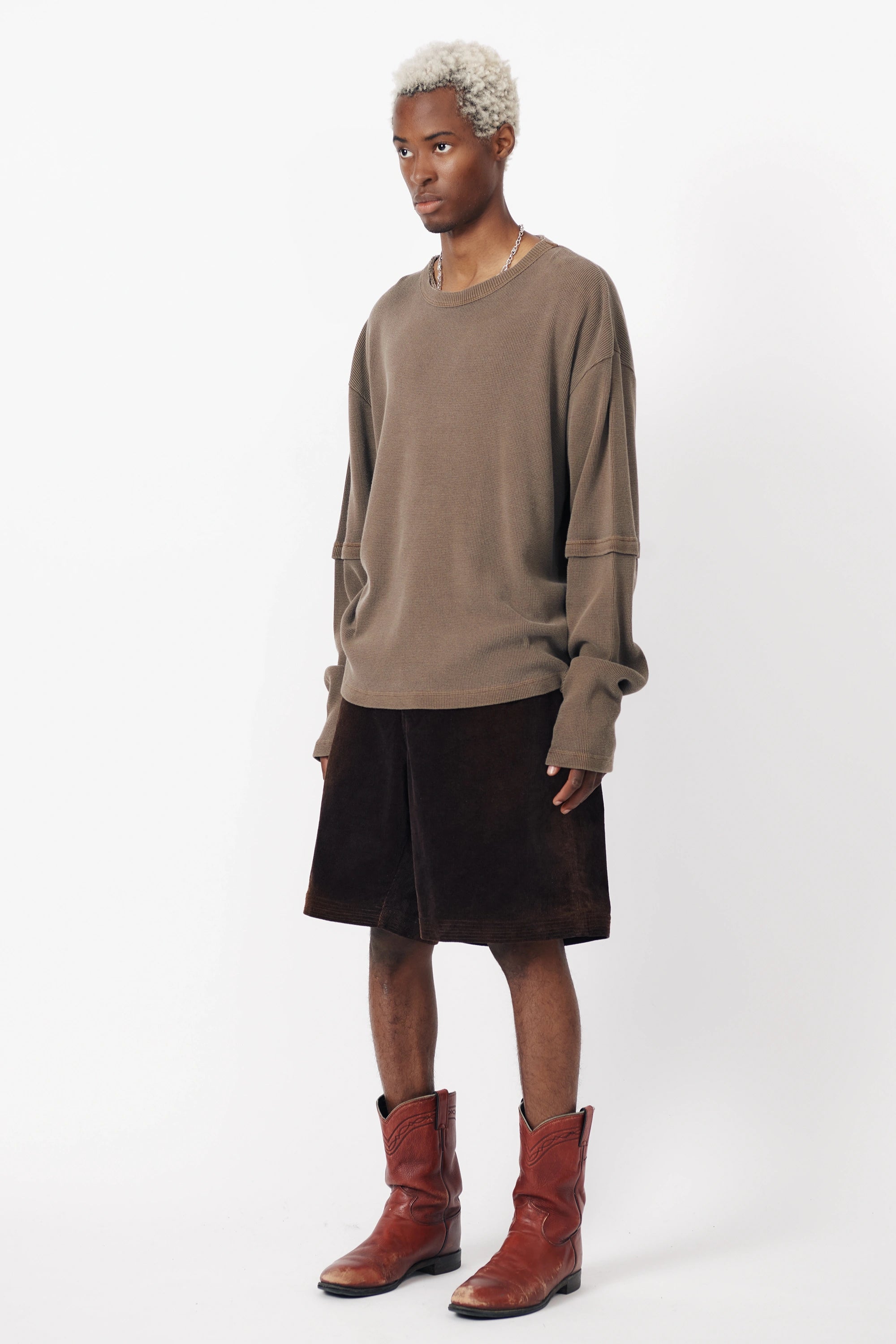 A person standing against a white background wearing a washed brown long-sleeve shirt and dark baggy shorts, with red boots.