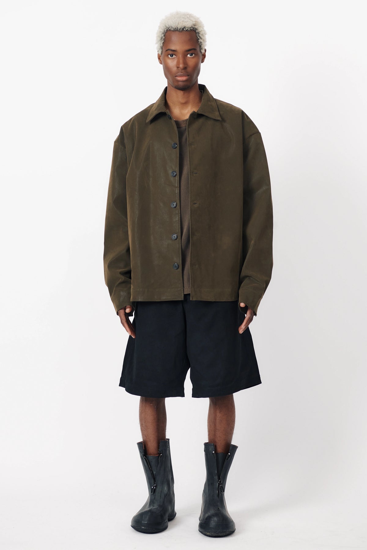 A person standing wearing black baggy shorts with a wide leg and an oversized green shirt with front pleats. Accessories include black boots.