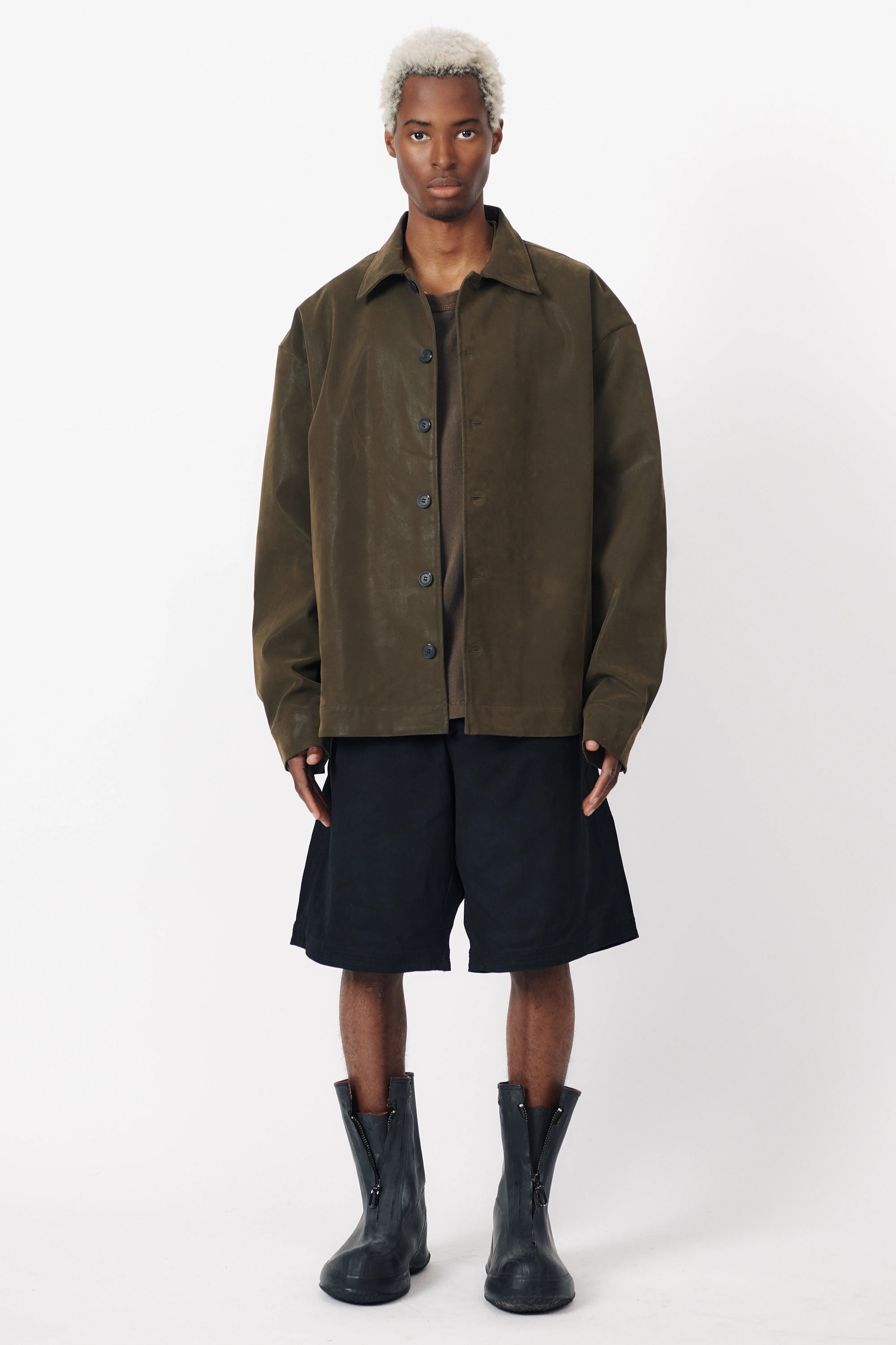 A person standing wearing black baggy shorts with a wide leg and an oversized green shirt with front pleats. Accessories include black boots.