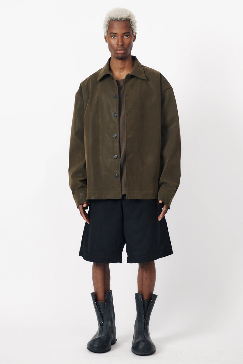 A person standing wearing black baggy shorts with a wide leg and an oversized green shirt with front pleats. Accessories include black boots.