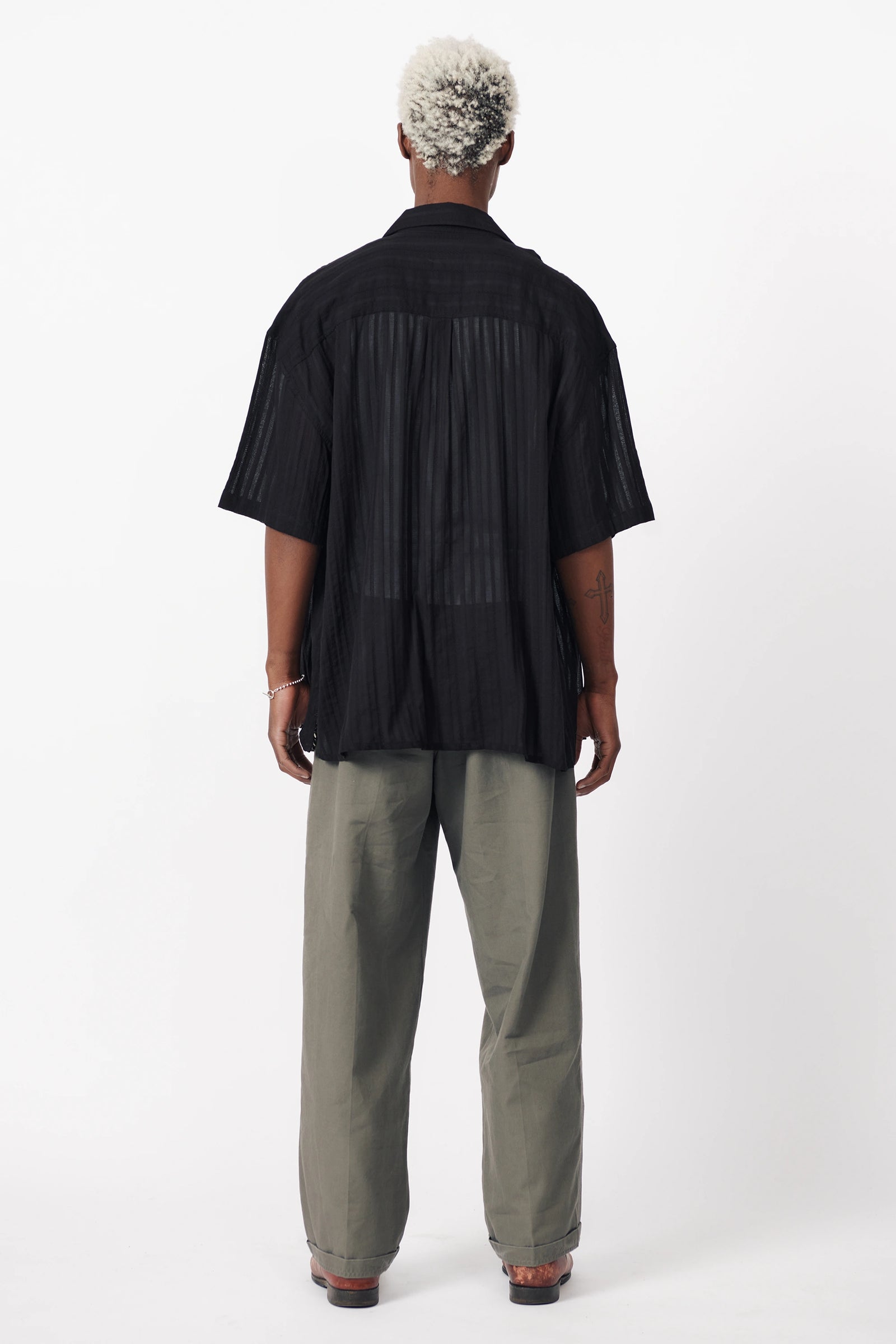 Rear view of a male model wearing a black, short-sleeved, button-up shirt paired with light grey trousers.
