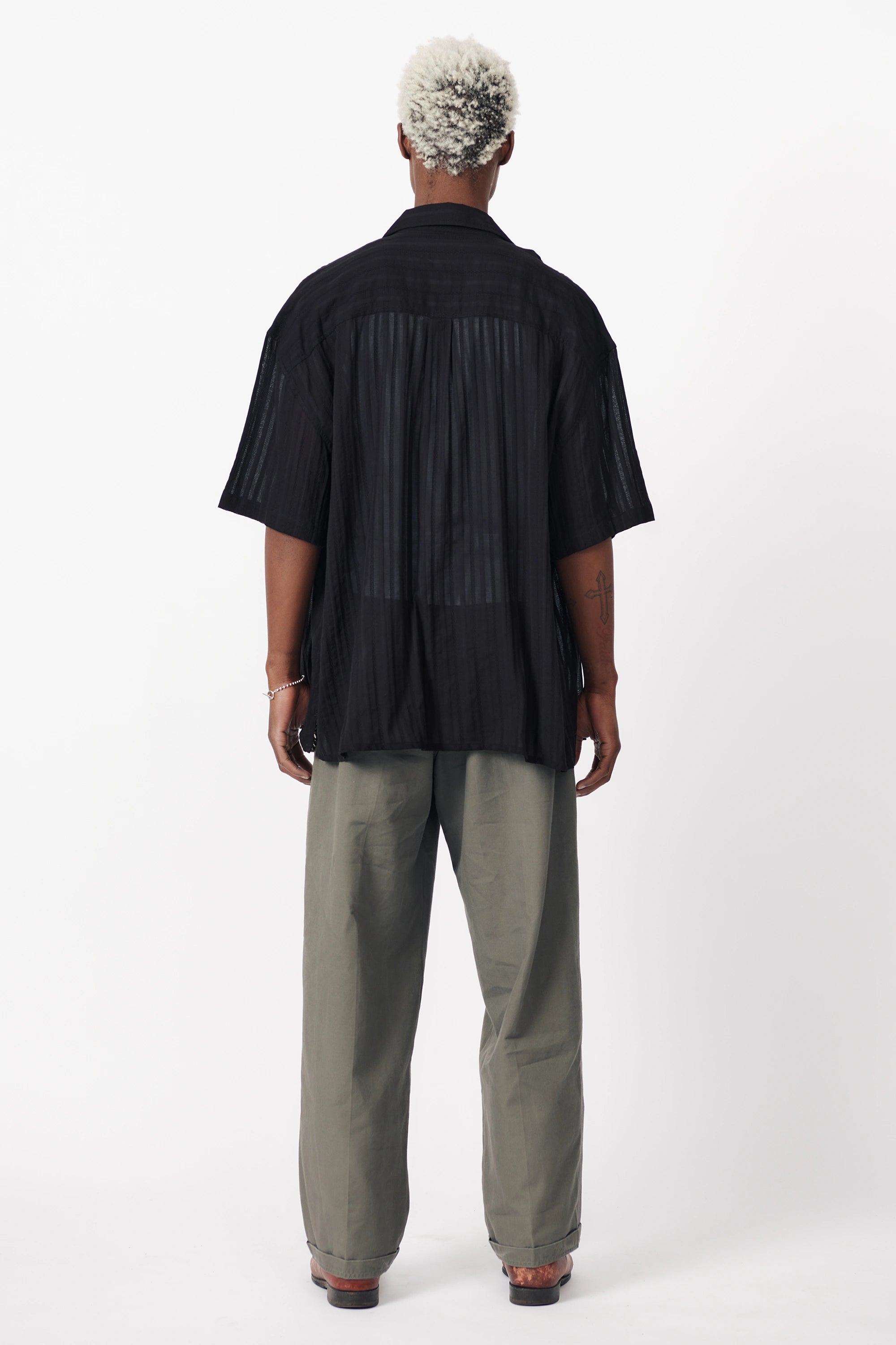 Rear view of a male model wearing a black, short-sleeved, button-up shirt paired with light grey trousers.