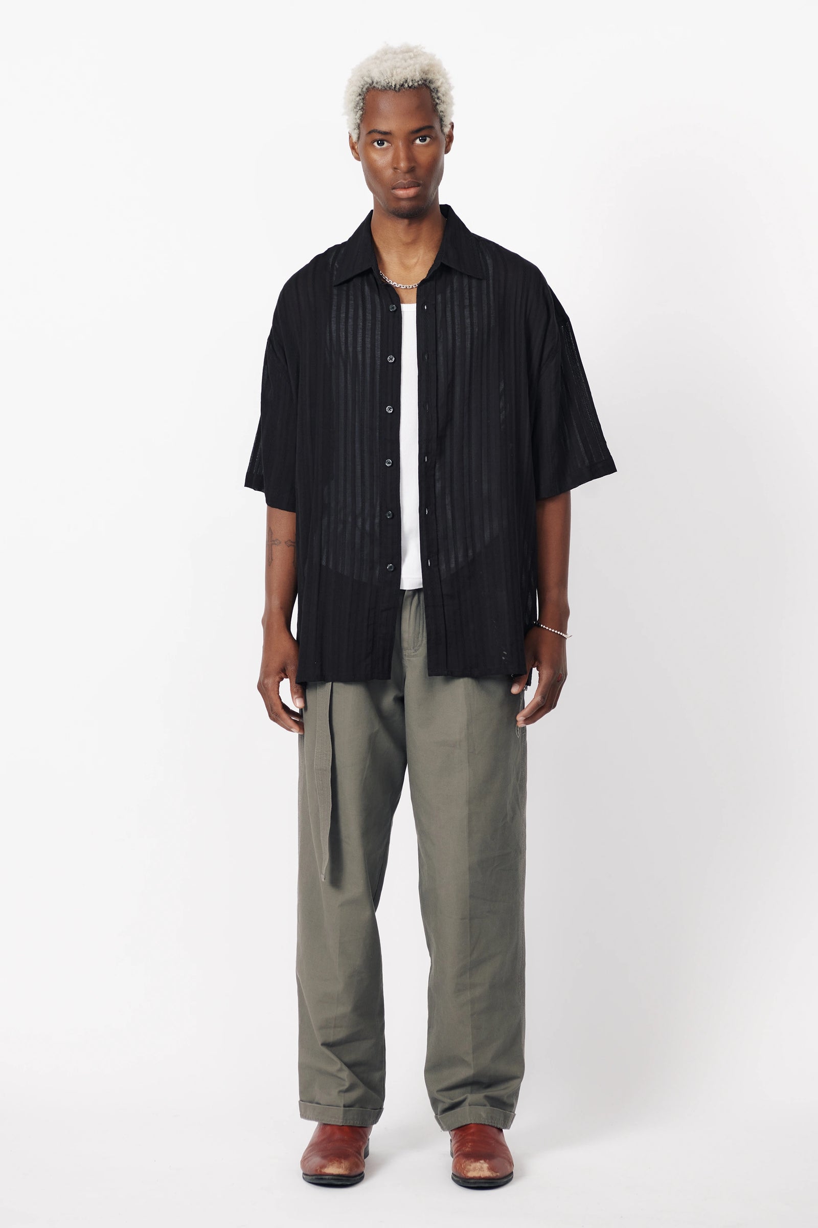 A man wearing a black, short-sleeved, button-up shirt with a geometric pattern and light grey trousers.