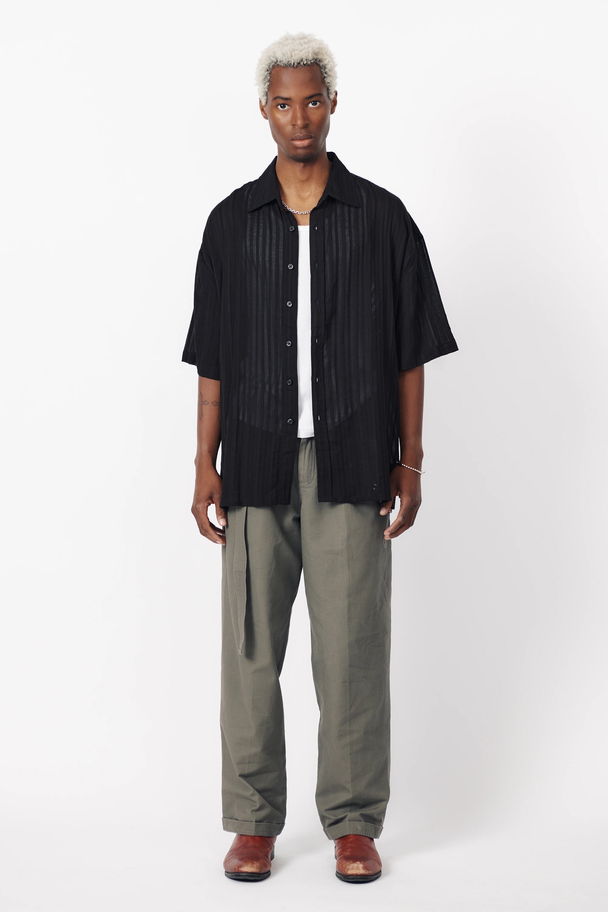 A man wearing a black, short-sleeved, button-up shirt with a geometric pattern and light grey trousers.