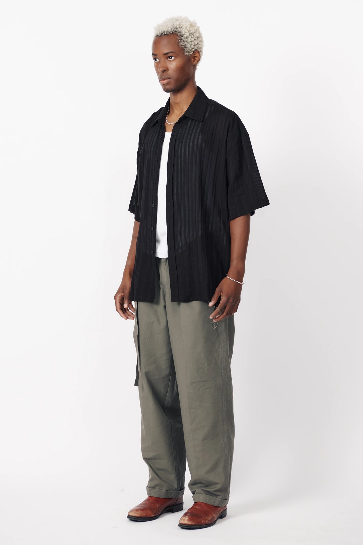 A male model facing off to the sid wearing a black, short-sleeved, button-up shirt with a textured pattern and light grey trousers