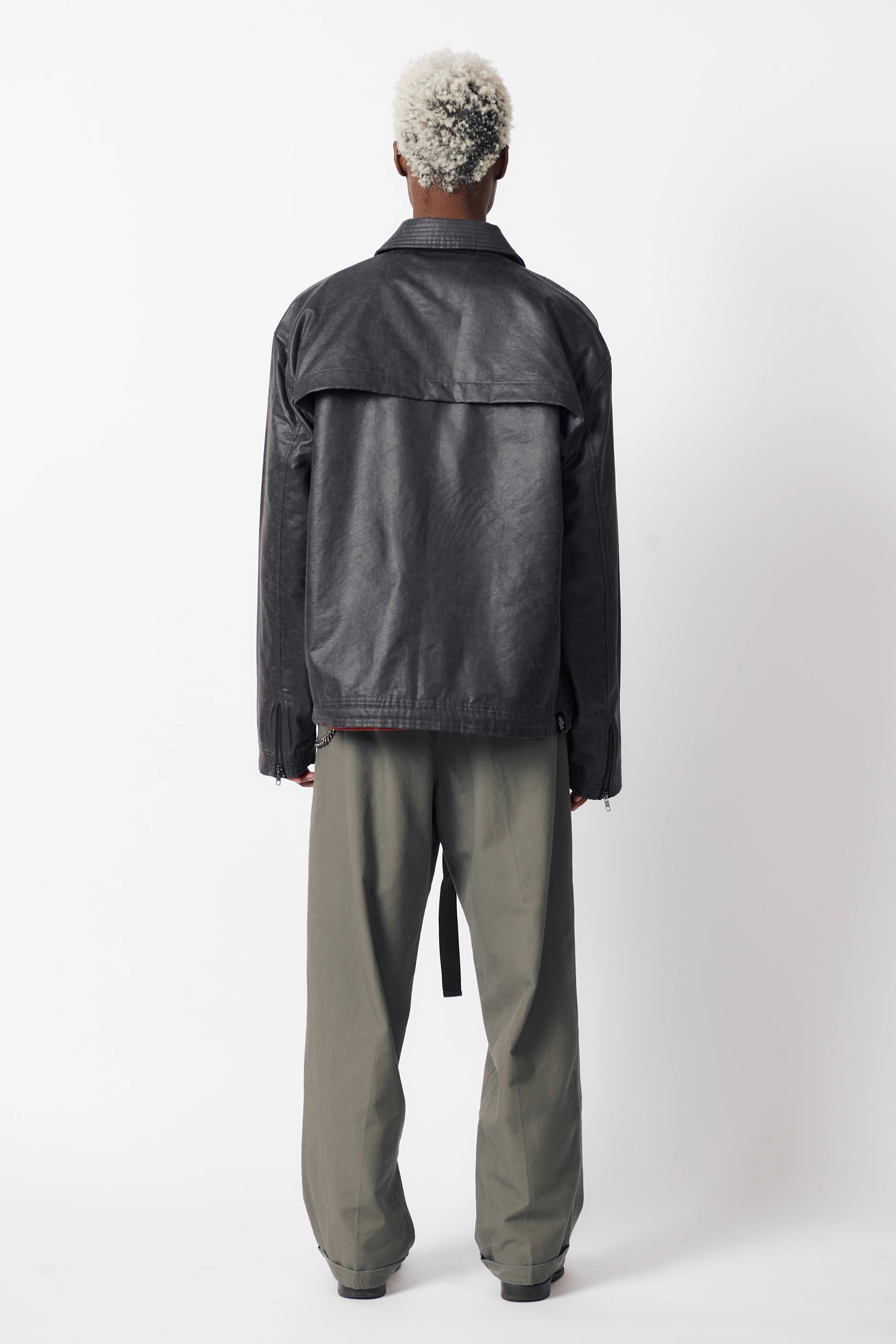 A male model with his back turned wearing a dark stone coloured coach jacket paired with grey trousers and standing against a white background