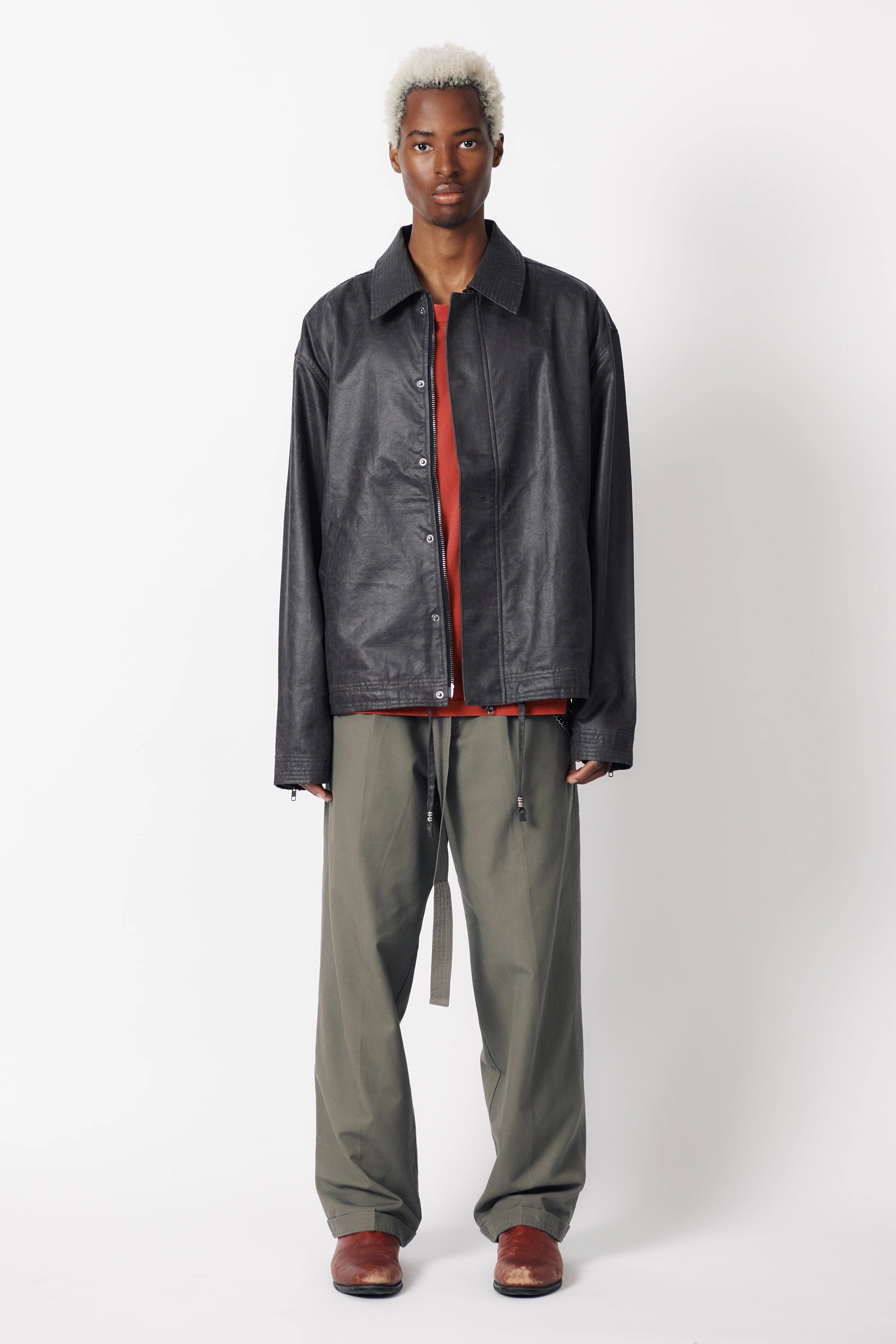 A male model wearing a dark stone coloured coach jacket with a collared neckline and long sleeves, paired with grey trousers and standing against a white background.