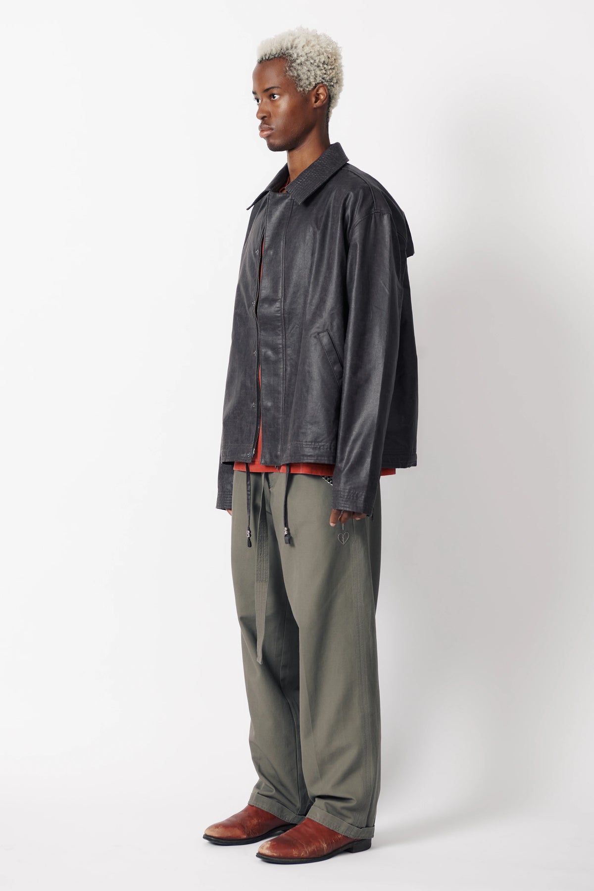 Side view of a man wearing a dark stone coloured coach jacket with a collared neckline and long sleeves paired with grey trousers.