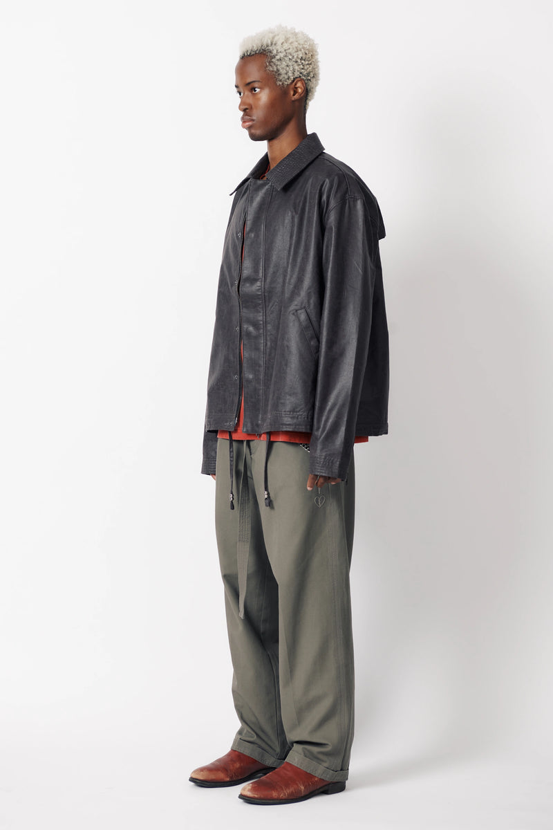 Side view of a man wearing a dark stone coloured coach jacket with a collared neckline and long sleeves paired with grey trousers.