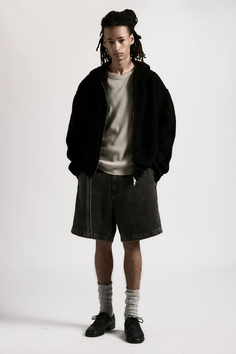 Person wearing a black jacket, grey shirt, black denim shorts and black shoes on a white background
