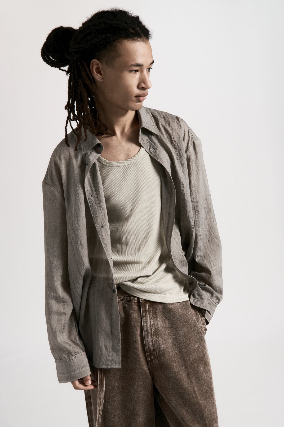 Person wearing a light grey shirt over a beige tank top with brown pants on a white background