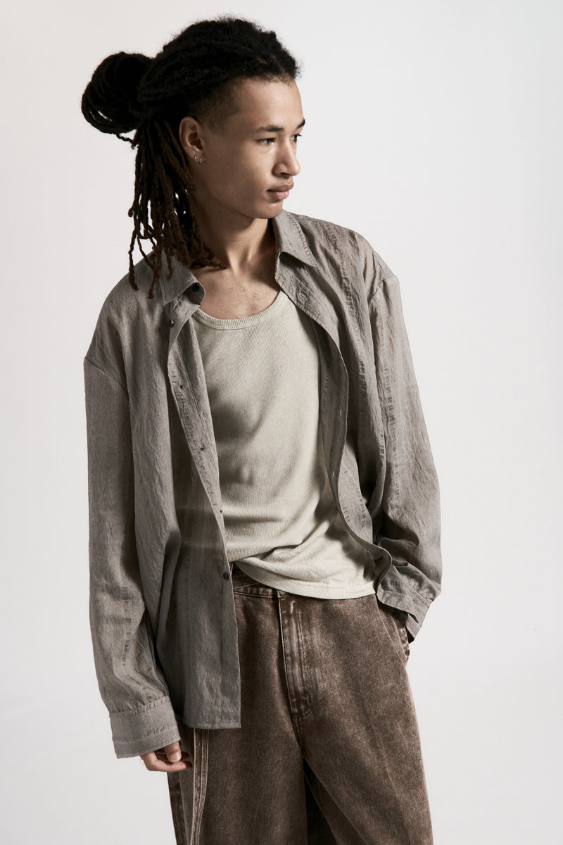 Person wearing a light grey shirt over a beige tank top with brown pants on a white background