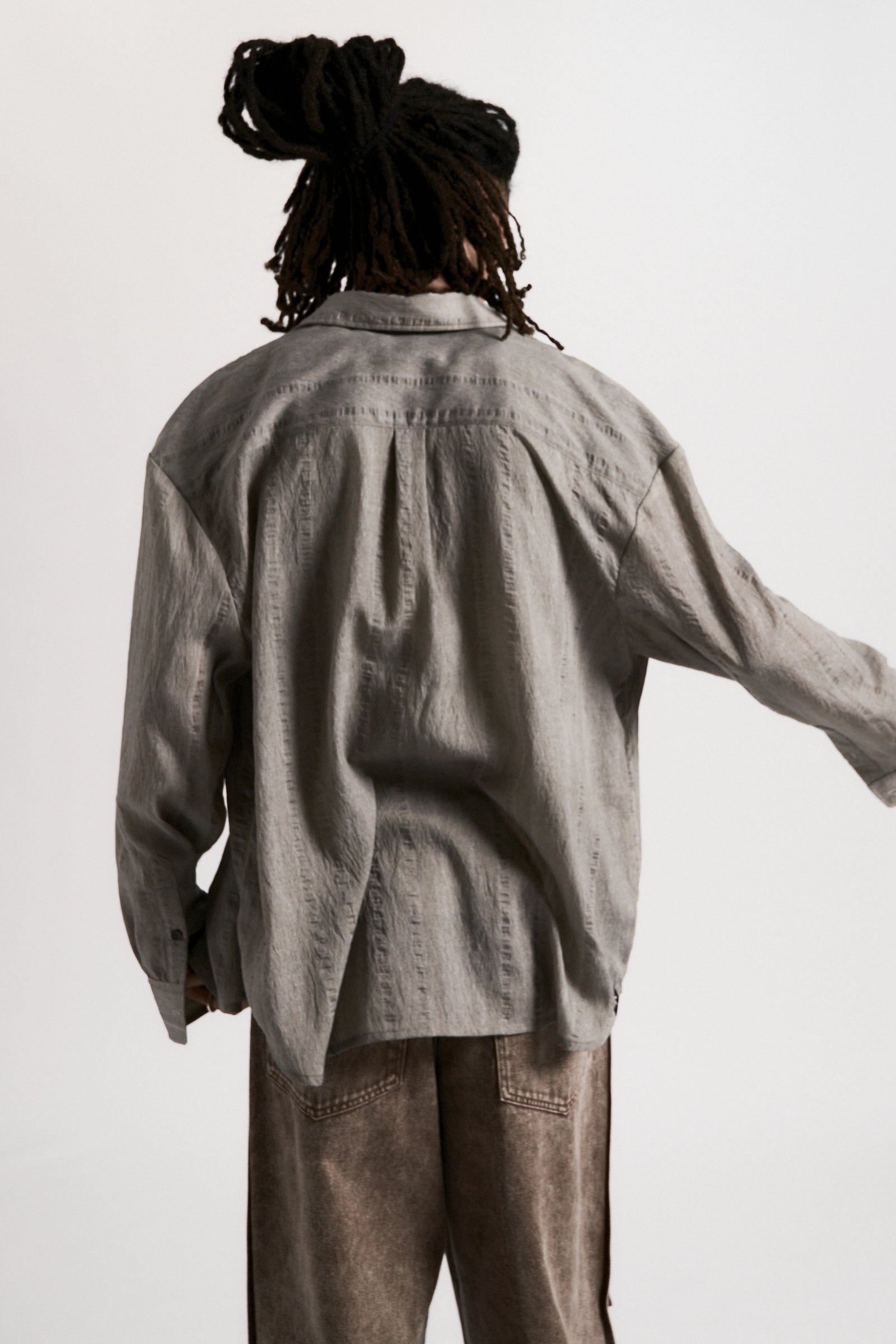 View from behind of a male model wearing a Sheer Grey Drop Long Sleeve Shirt and brown pants against a white background