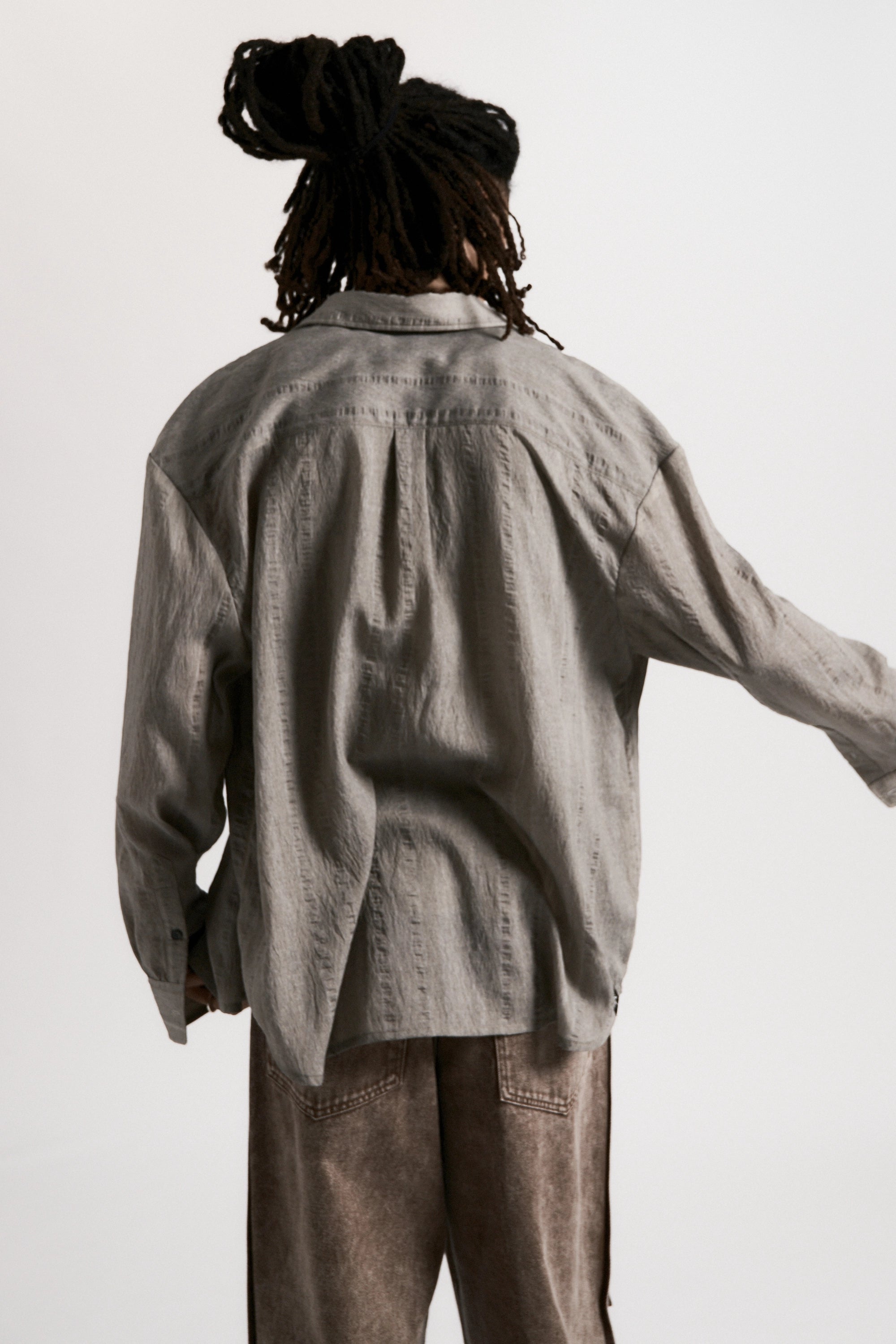 View from behind of a male model wearing a Sheer Grey Drop Long Sleeve Shirt and brown pants against a white background