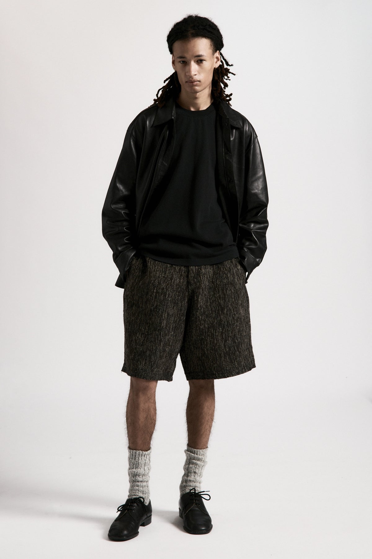 Person wearing a black leather overshirt, black t-shirt underneath and grey shorts on a white background