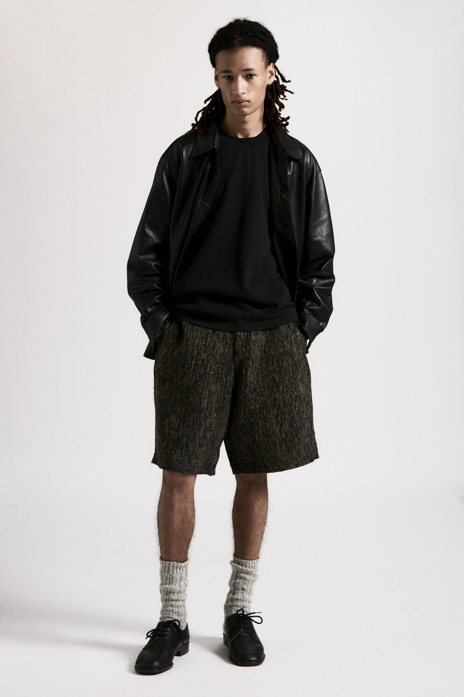Person wearing a black leather overshirt, black t-shirt underneath and grey shorts on a white background