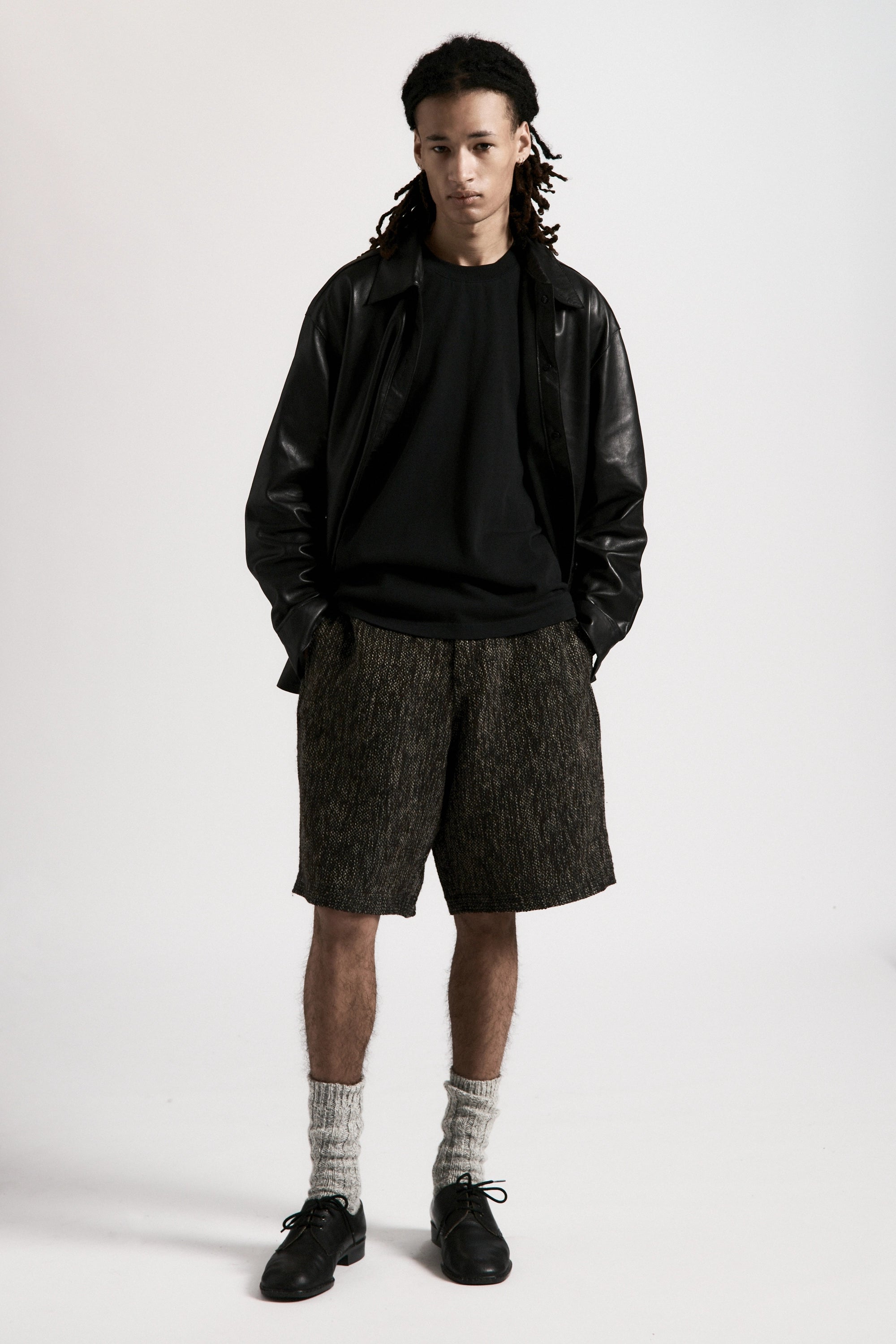 Person wearing a black leather overshirt, black t-shirt underneath and grey shorts on a white background