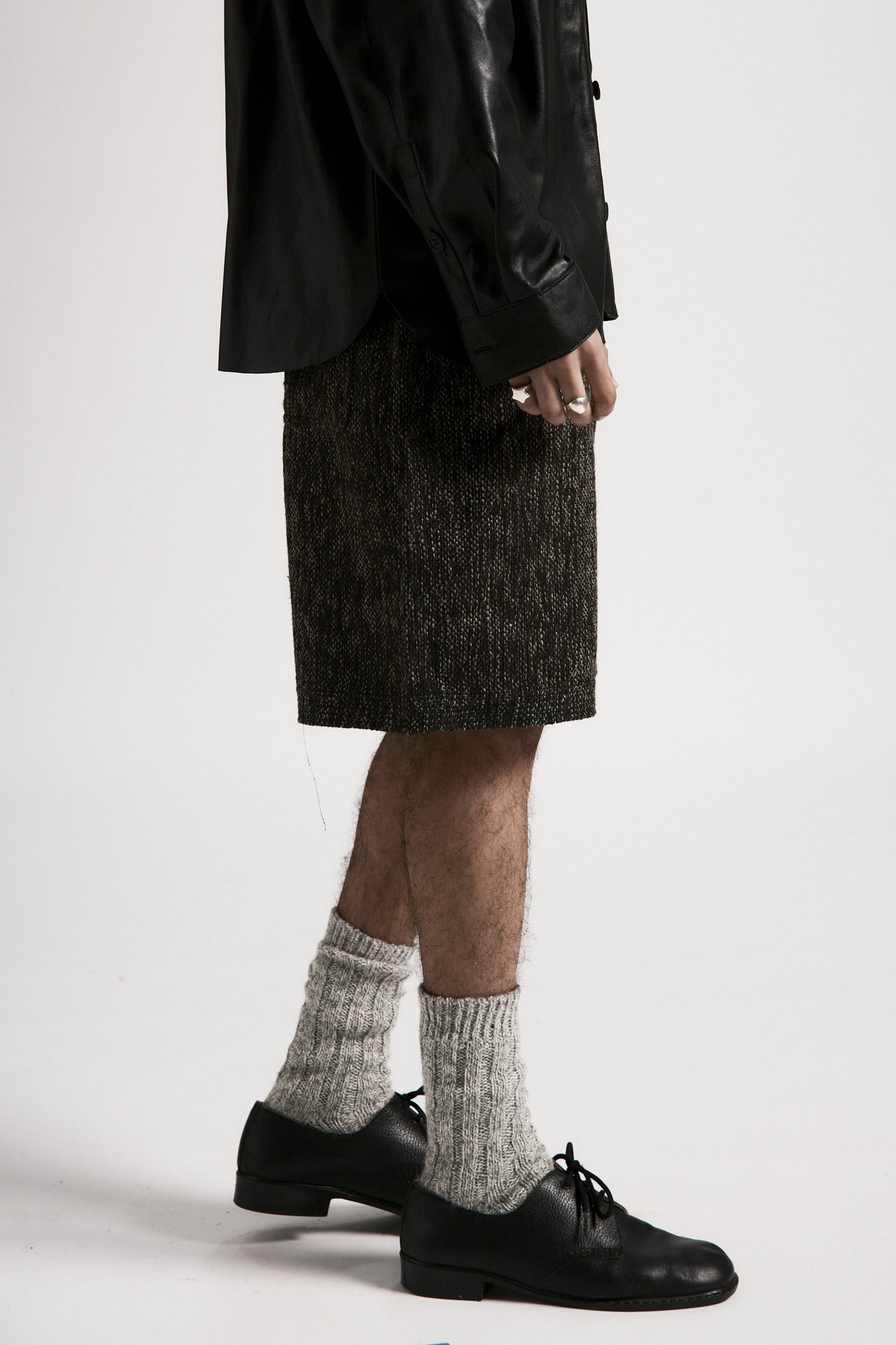 Person wearing a dark leather overshirt, grey shirt and black shoes with white socks on a plain background