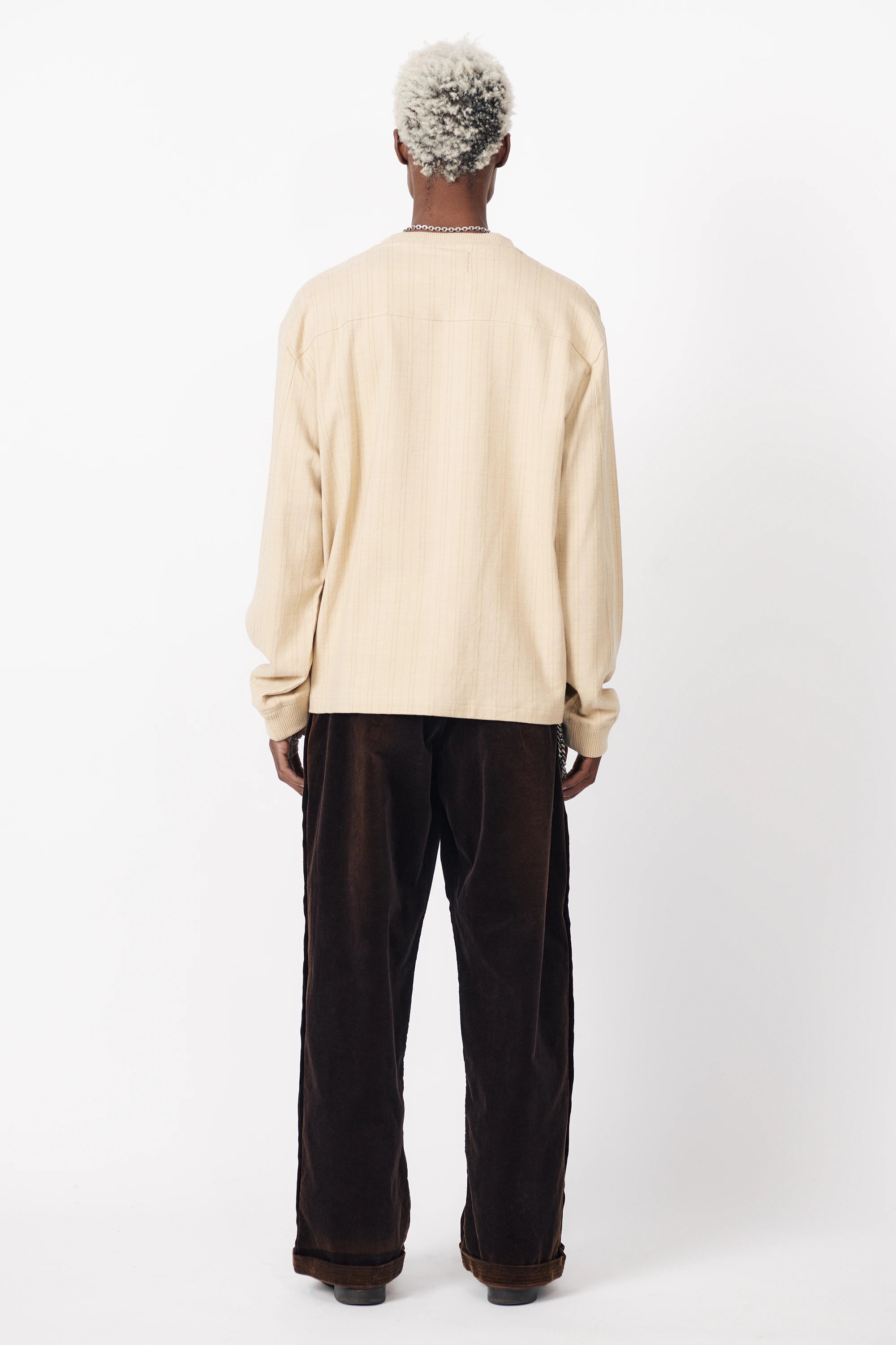 View from behind a person standing against a white background wearing a beige v-neck football shirt with tan stripes and long sleeves and paired with dark pants.
