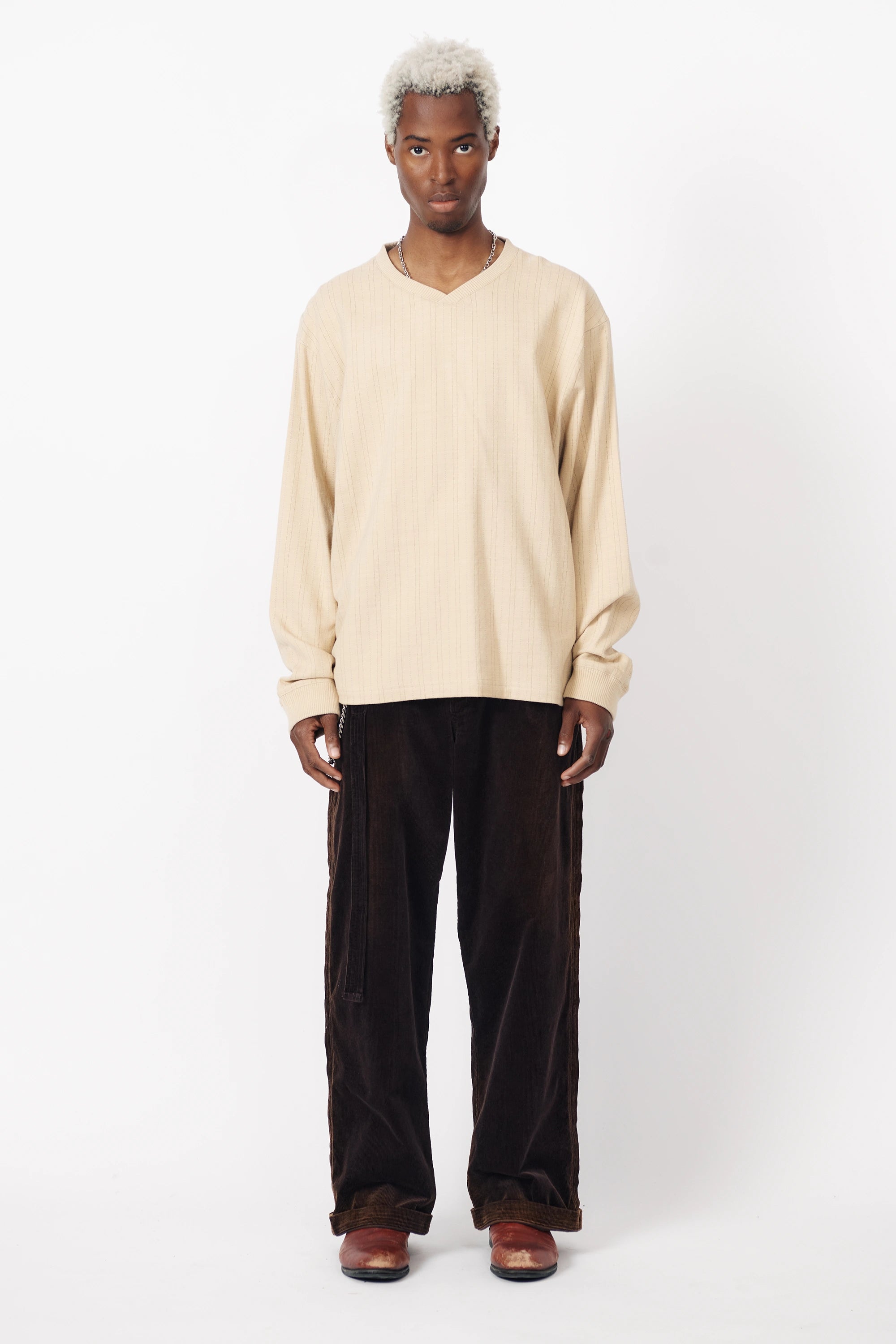 A person standing against a white background wearing a beige v-neck football shirt with tan stripes and long sleeves, paired with dark pants.