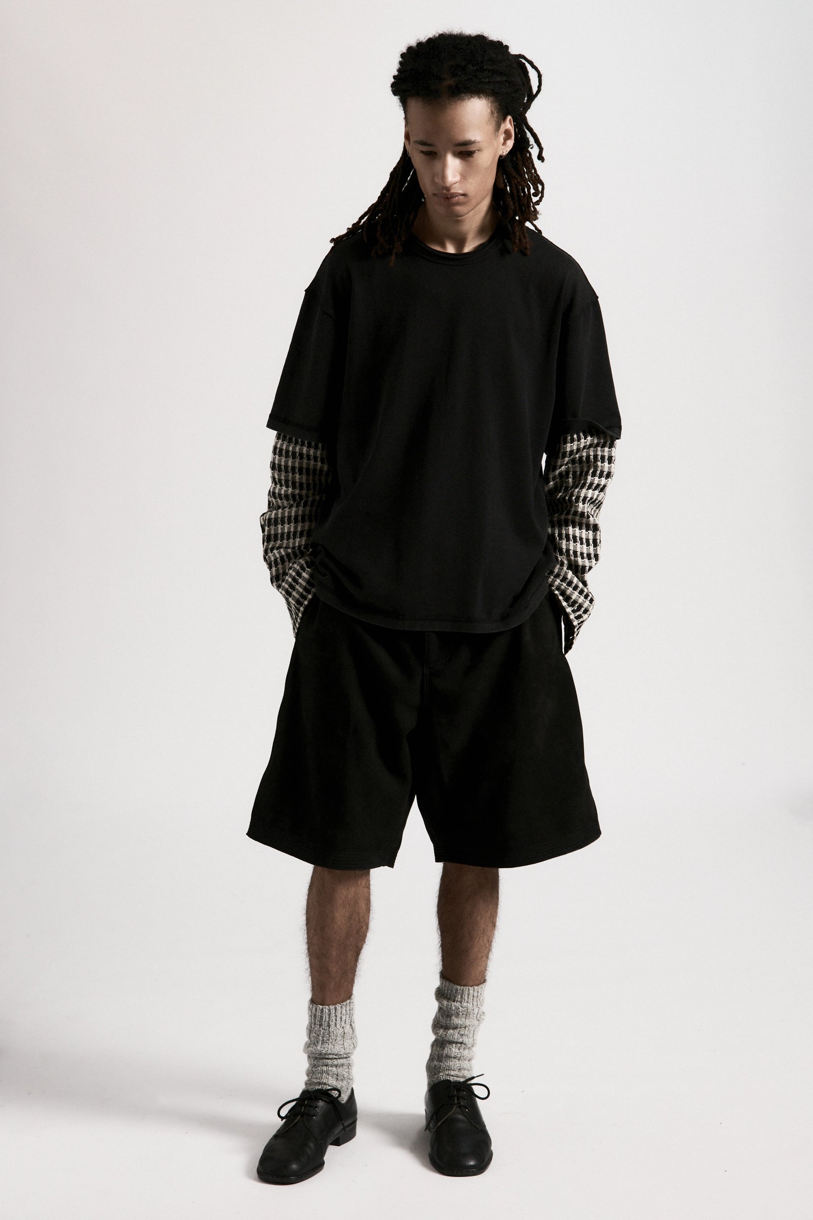 Person wearing a black shirt with checkered sleeves and black hang shorts on a white background