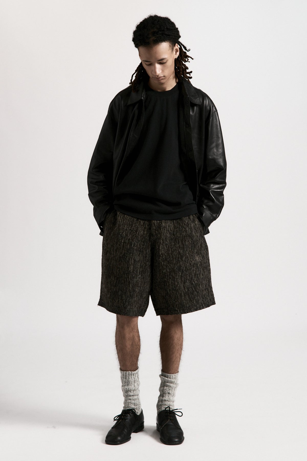 Person wearing a black leather jacket, black shirt and grey fossil hang shorts on a white background