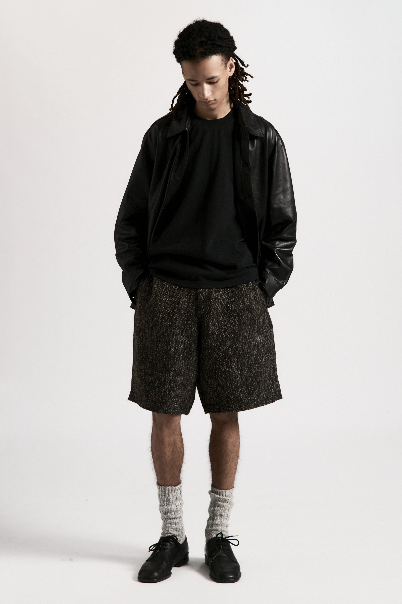 Person wearing a black leather jacket, black shirt and grey fossil hang shorts on a white background