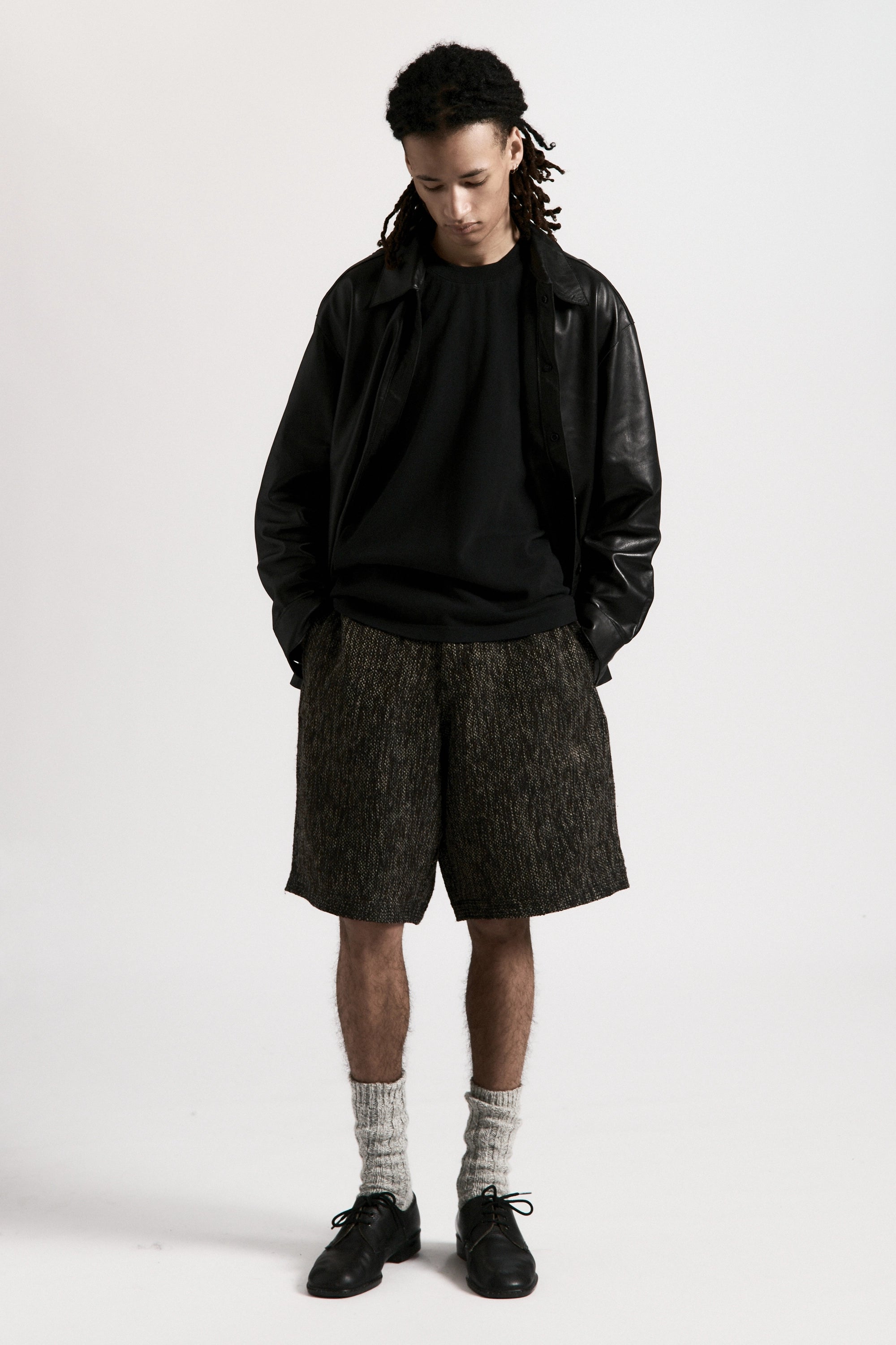 Person wearing a black leather jacket, black shirt and grey fossil hang shorts on a white background