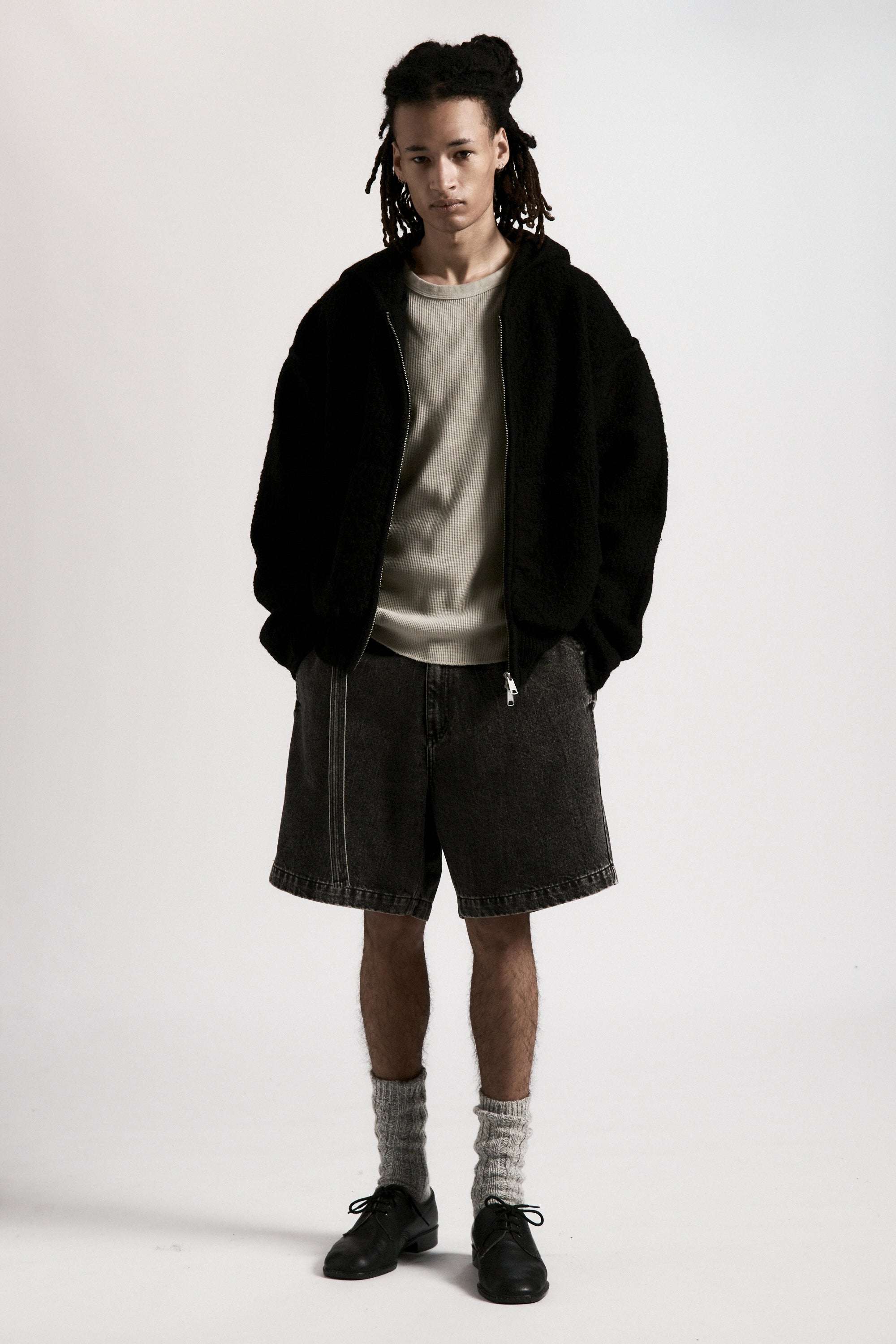 Person wearing a black boiled wool zipped hoodie, grey shirt, black shorts and black shoes on a white background