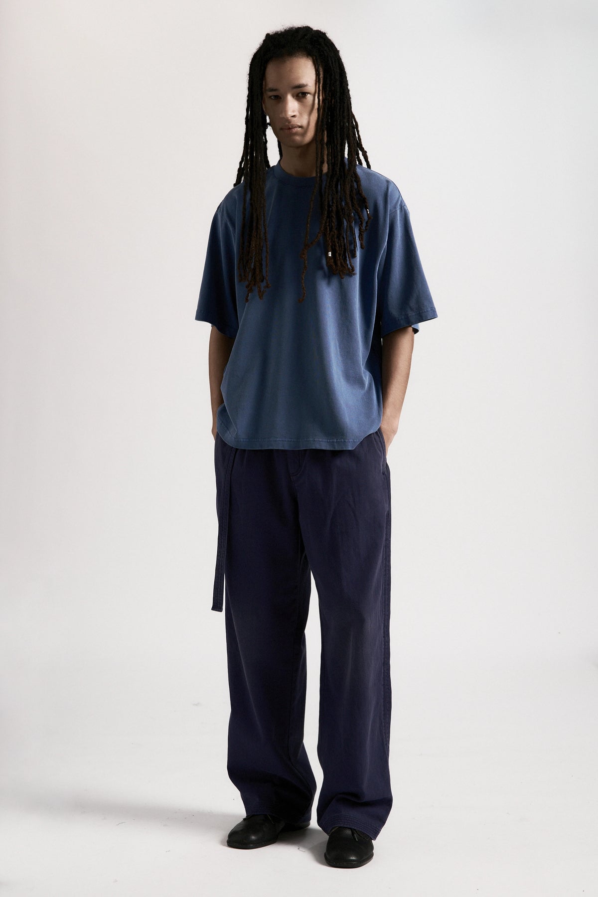 A person standing against a white background wearing a white t-shirt and pleated work pants in a navy blue colour.
