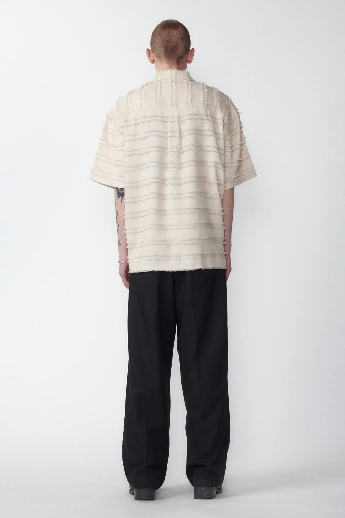View from the back of a male model wearing a boxy short-sleeved shirt with a striped pattern and black pants.