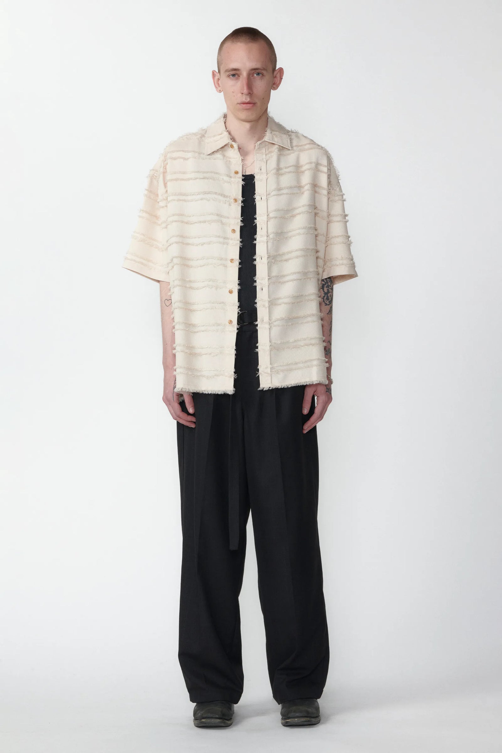 A person standing against a white background wearing a boxy short-sleeved shirt with a striped pattern and raw hem, paired with black pants.