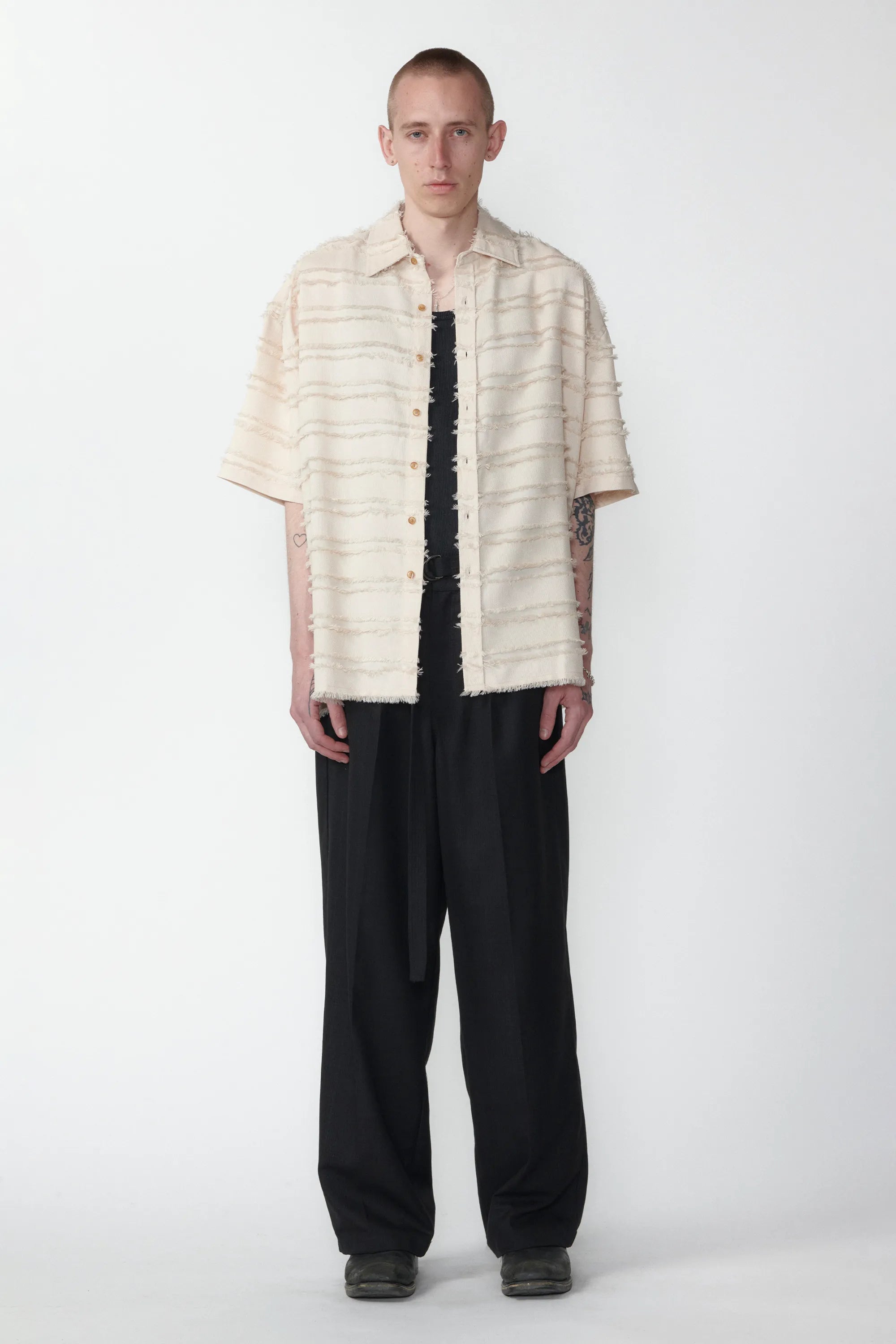 A person standing against a white background wearing a boxy short-sleeved shirt with a striped pattern and raw hem, paired with black pants.