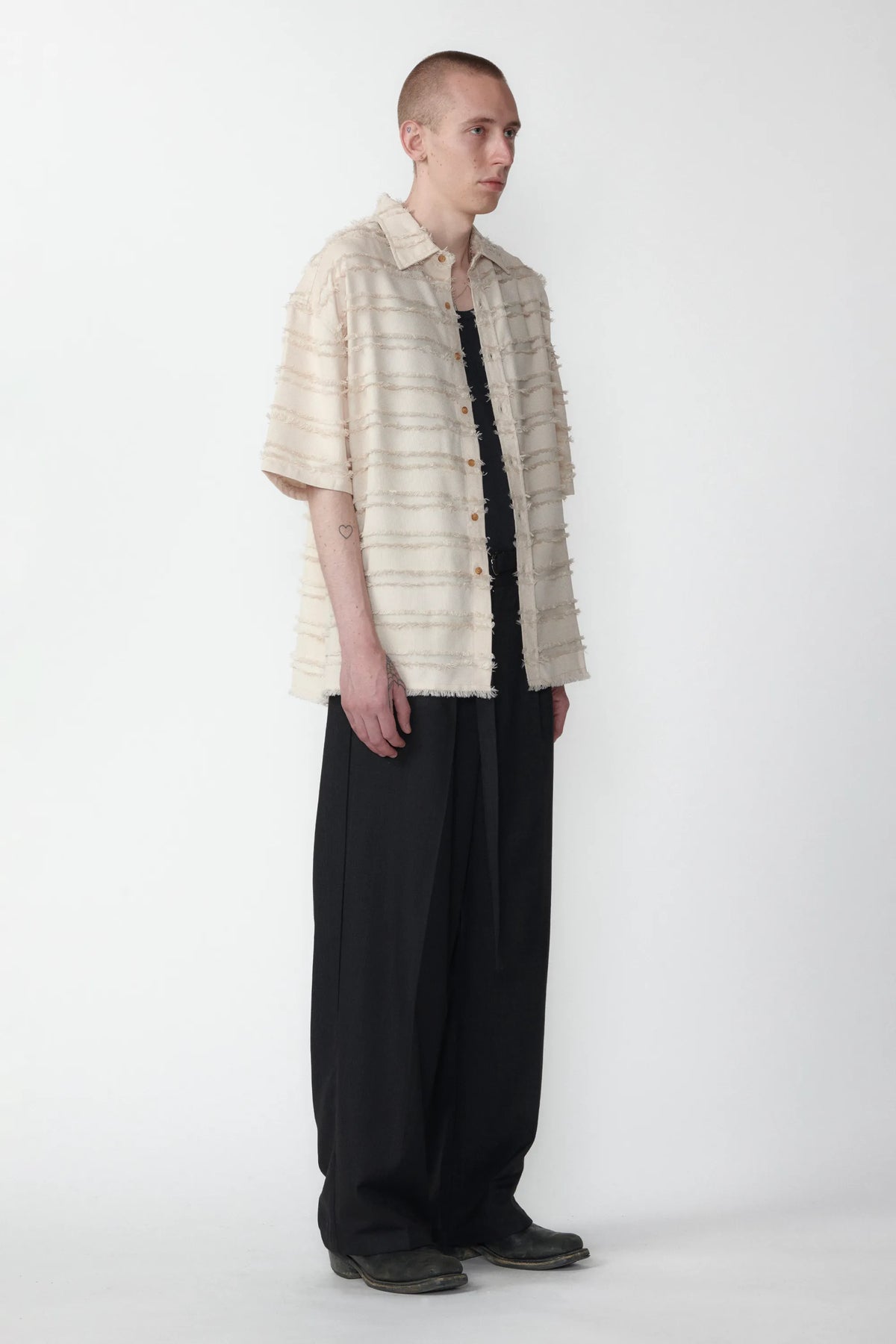 A male model standing against a white background facing off in one direction wearing a boxy short-sleeved shirt with a striped pattern paired with black pants.