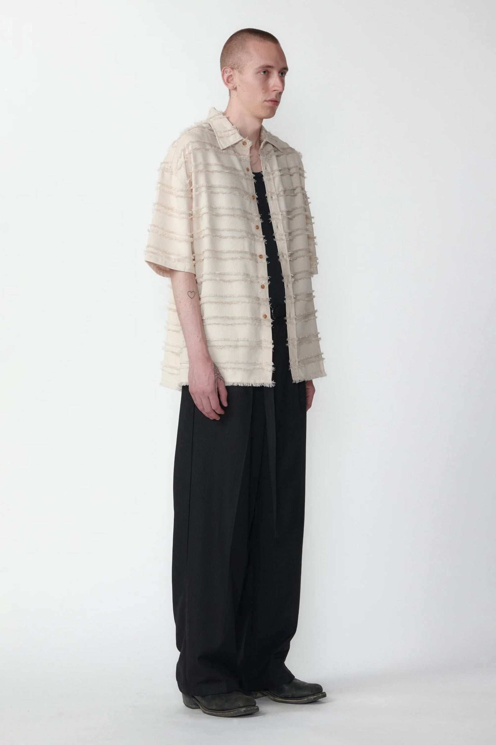 A male model standing against a white background facing off in one direction wearing a boxy short-sleeved shirt with a striped pattern paired with black pants.