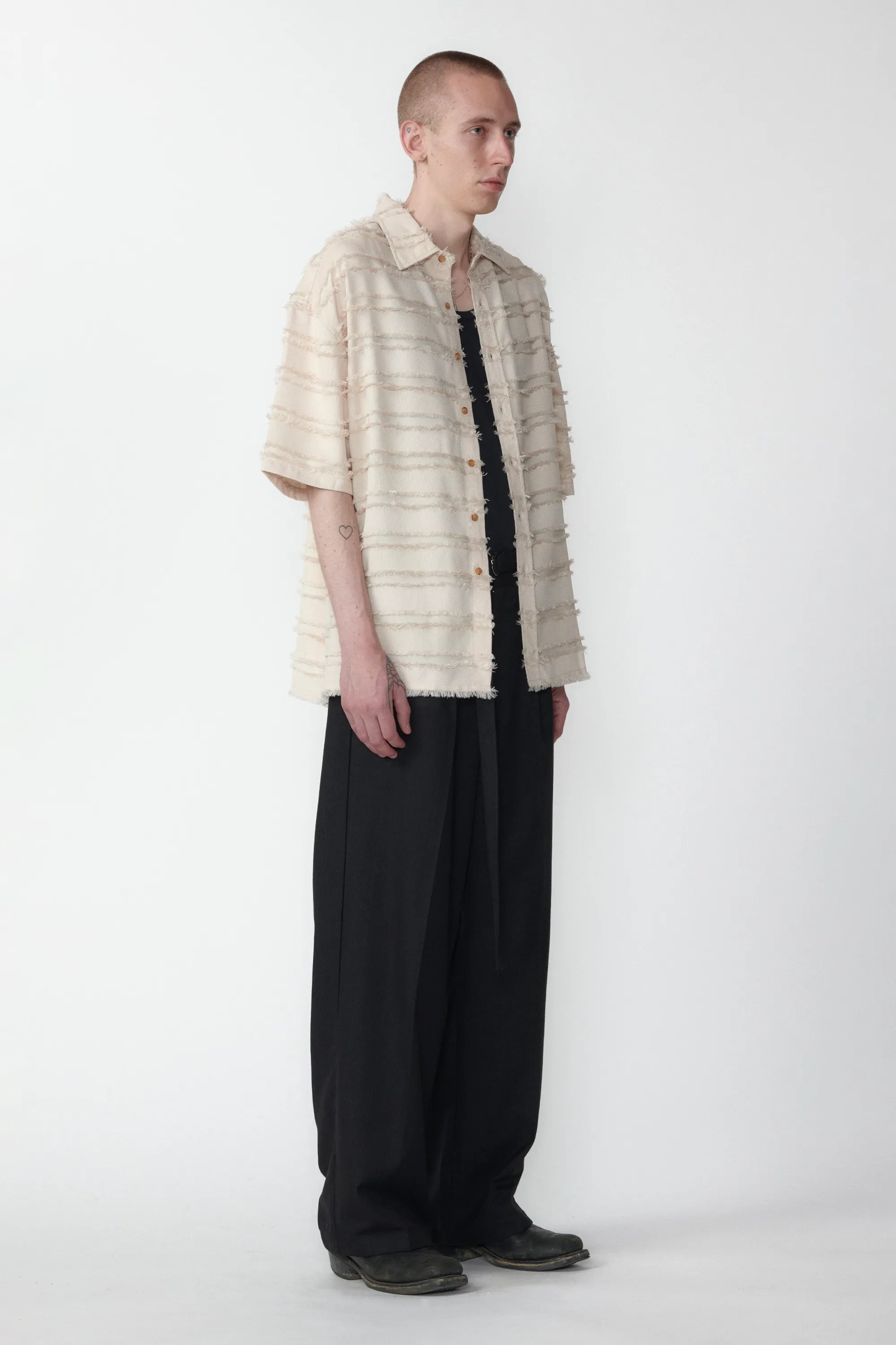 A male model standing against a white background facing off in one direction wearing a boxy short-sleeved shirt with a striped pattern paired with black pants.