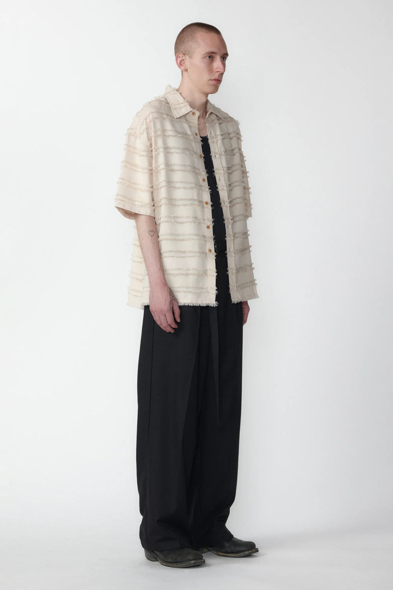 A male model standing against a white background facing off in one direction wearing a boxy short-sleeved shirt with a striped pattern paired with black pants.