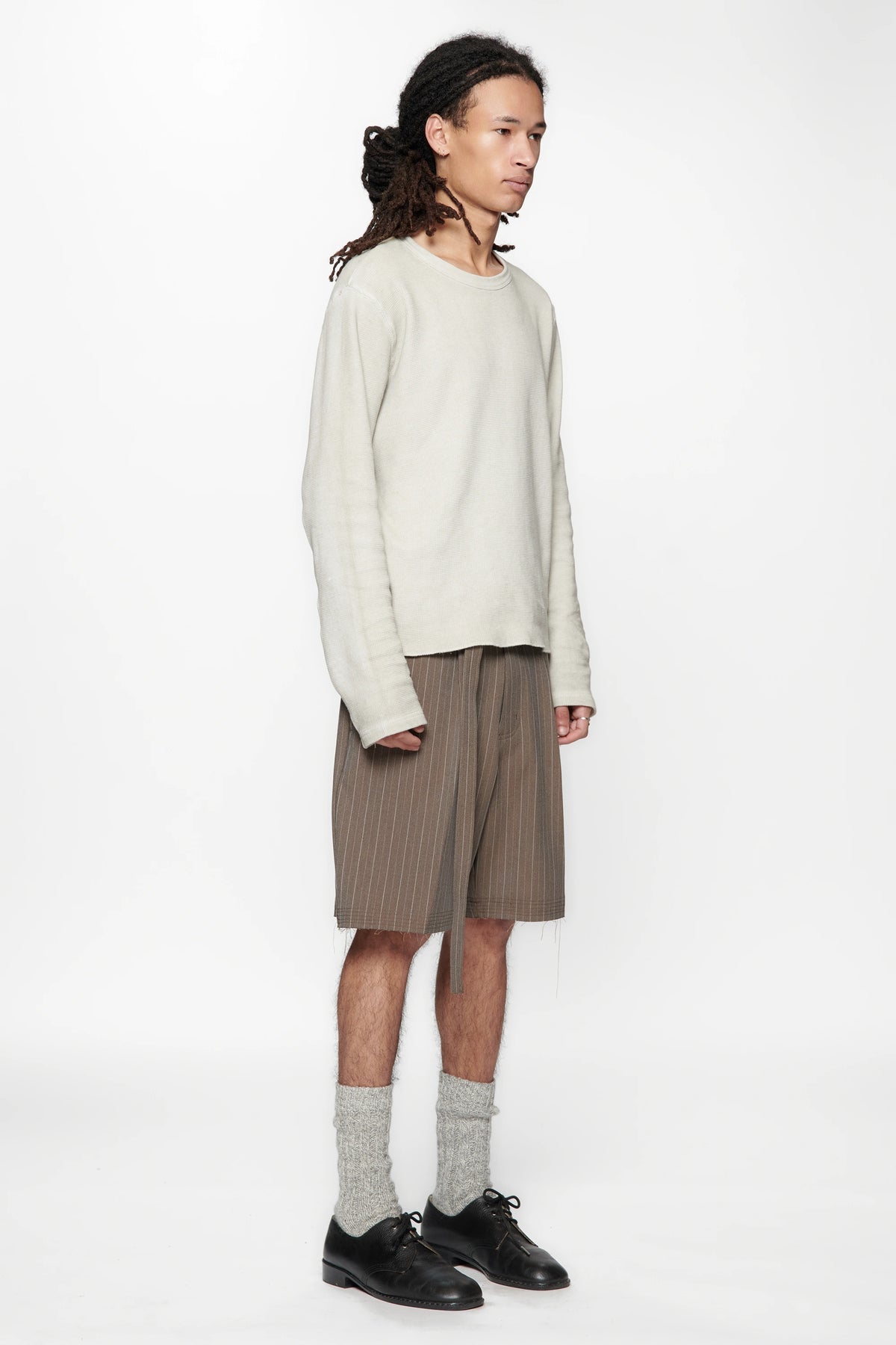 A male model wearing a light-coloured sweater and brown pinstripe cut shorts on a white background