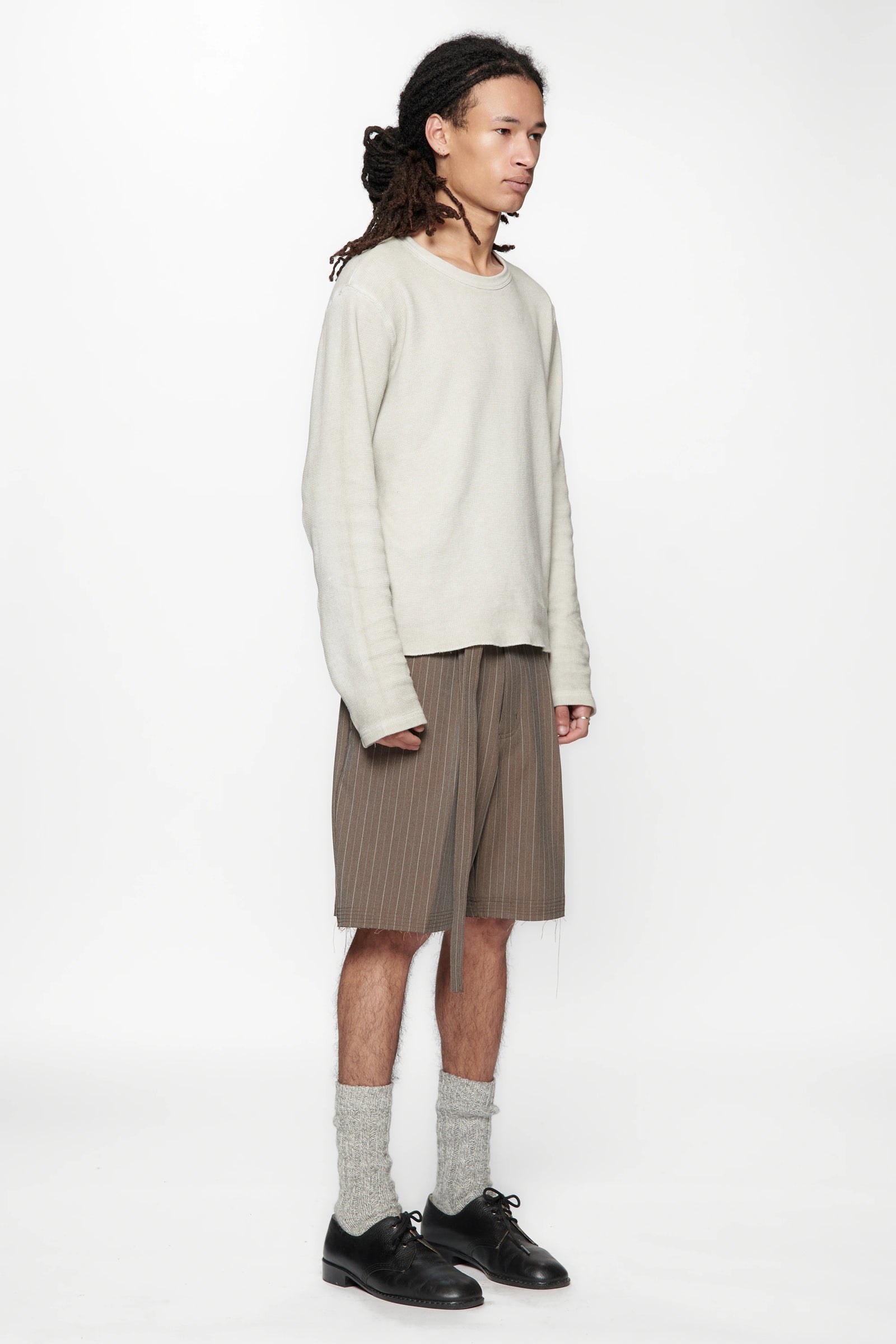 A male model wearing a light-coloured sweater and brown pinstripe cut shorts on a white background