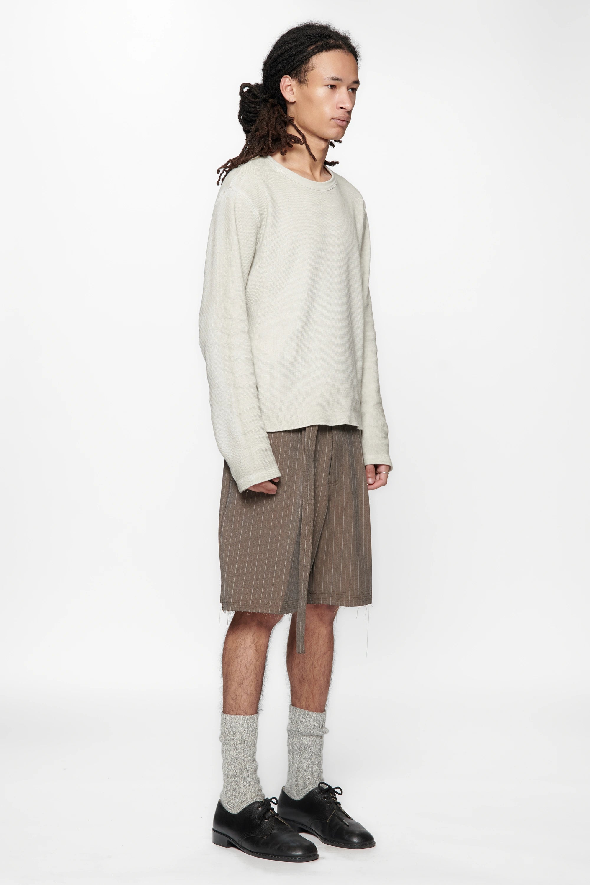 A male model wearing a light-coloured sweater and brown pinstripe cut shorts on a white background