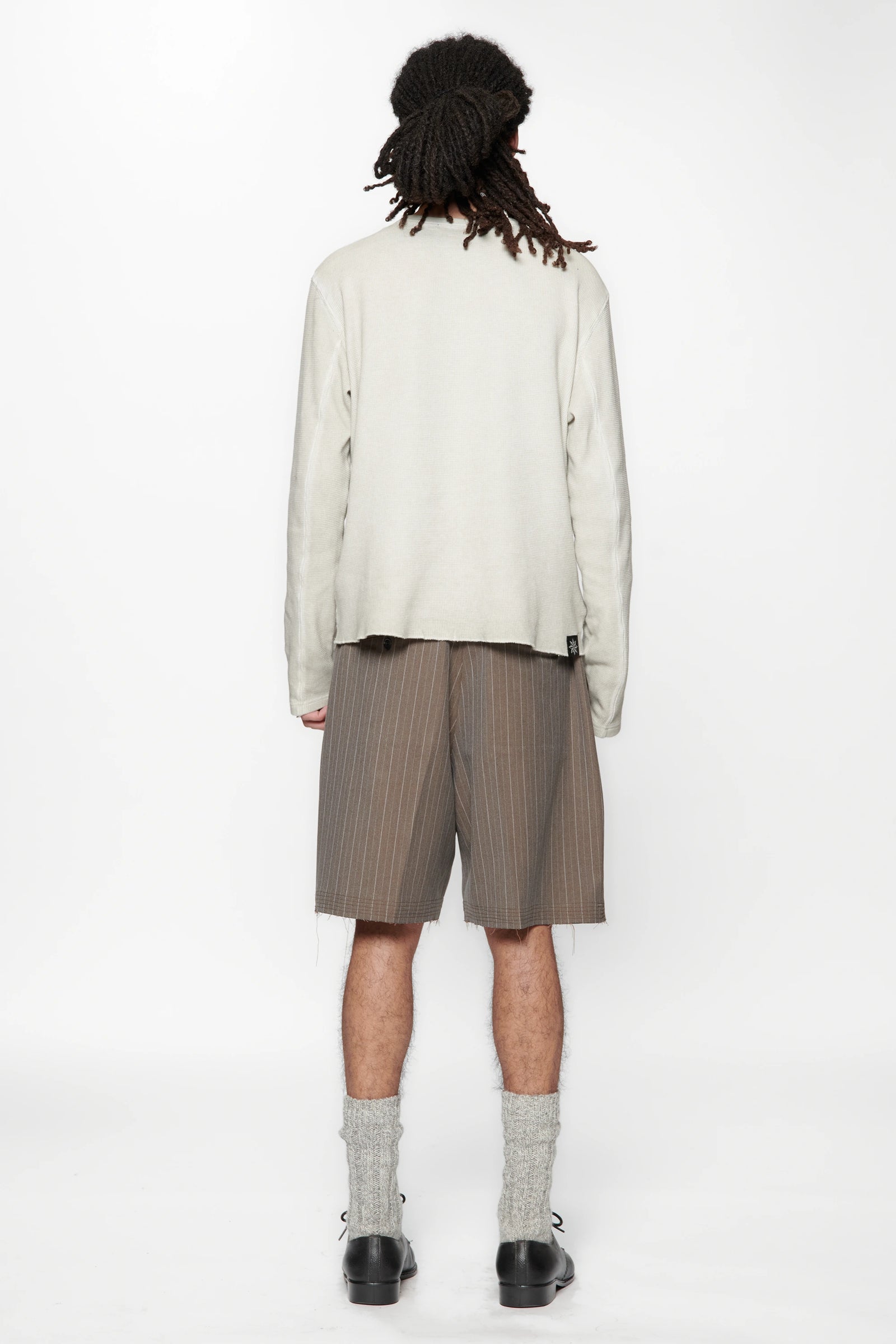 A male model with his back turned wearing a light-coloured shirt and cut pinstripe brown shorts on a white background