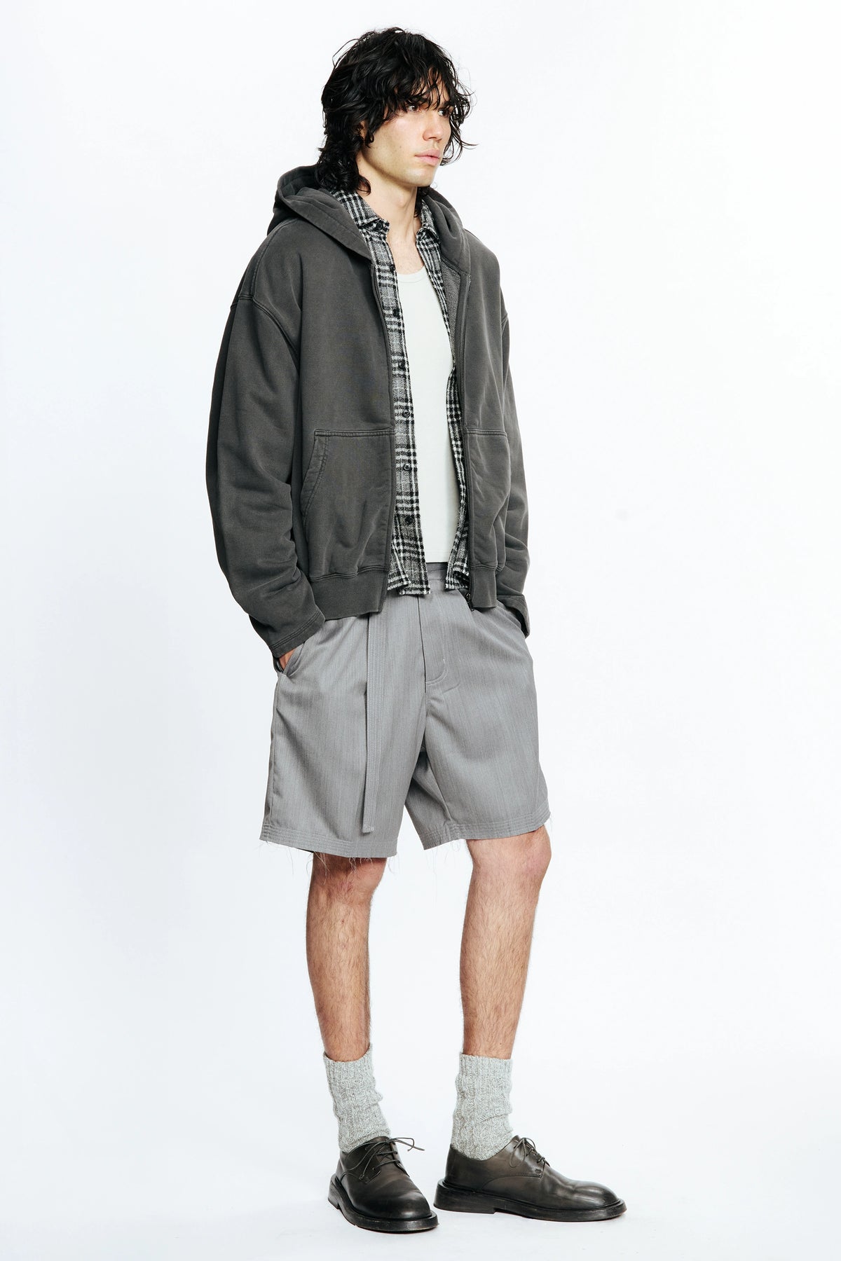 View from the side of a person wearing a grey jacket, plaid shirt and grey cut shorts on a white background