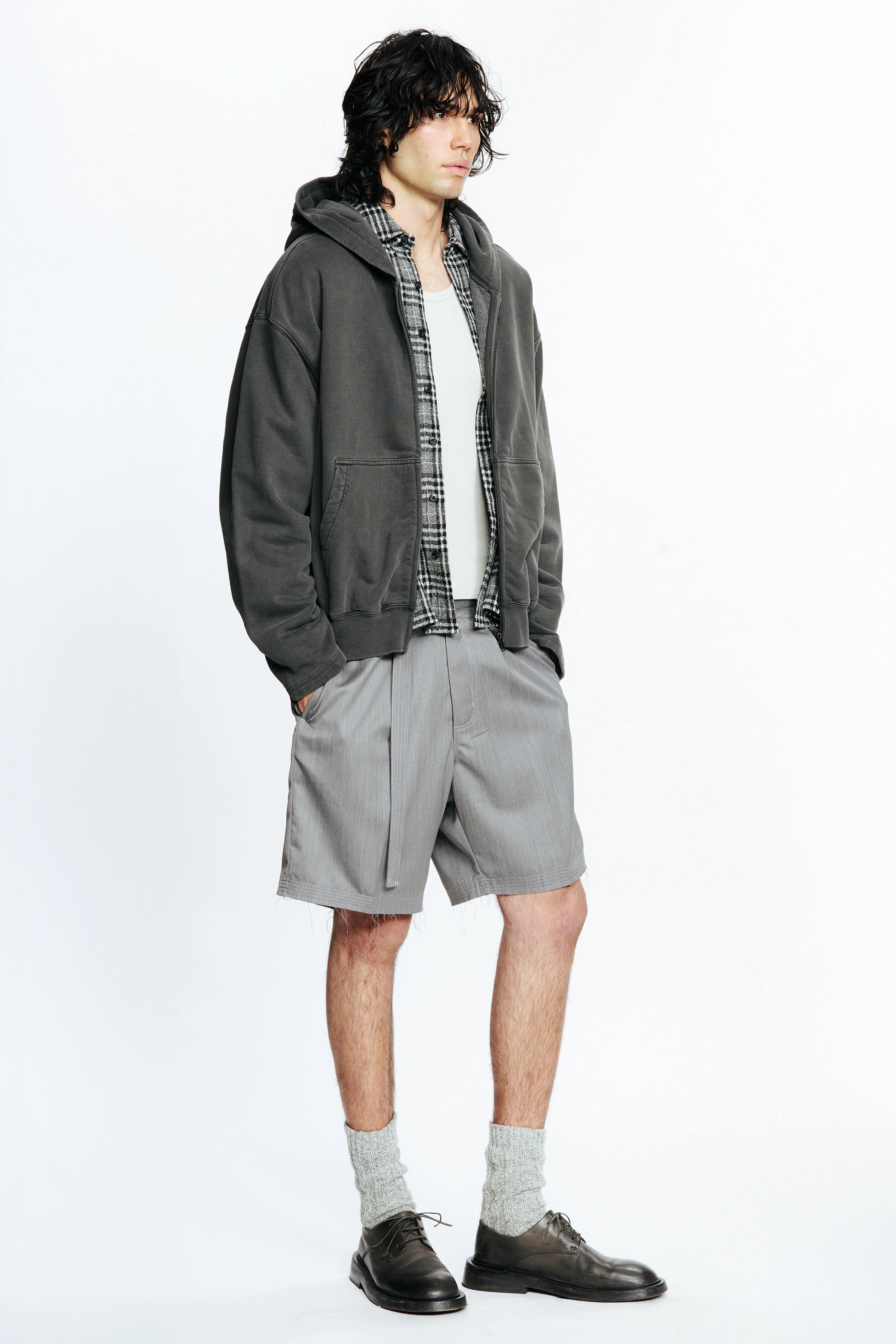 View from the side of a person wearing a grey jacket, plaid shirt and grey cut shorts on a white background