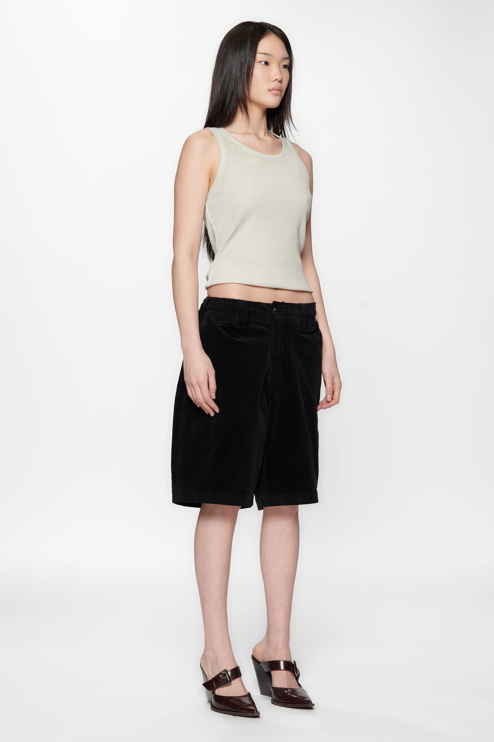 Woman facing t o the side wearing a beige sleeveless top and Vintage Black Corduroy Hang Shorts on a white background